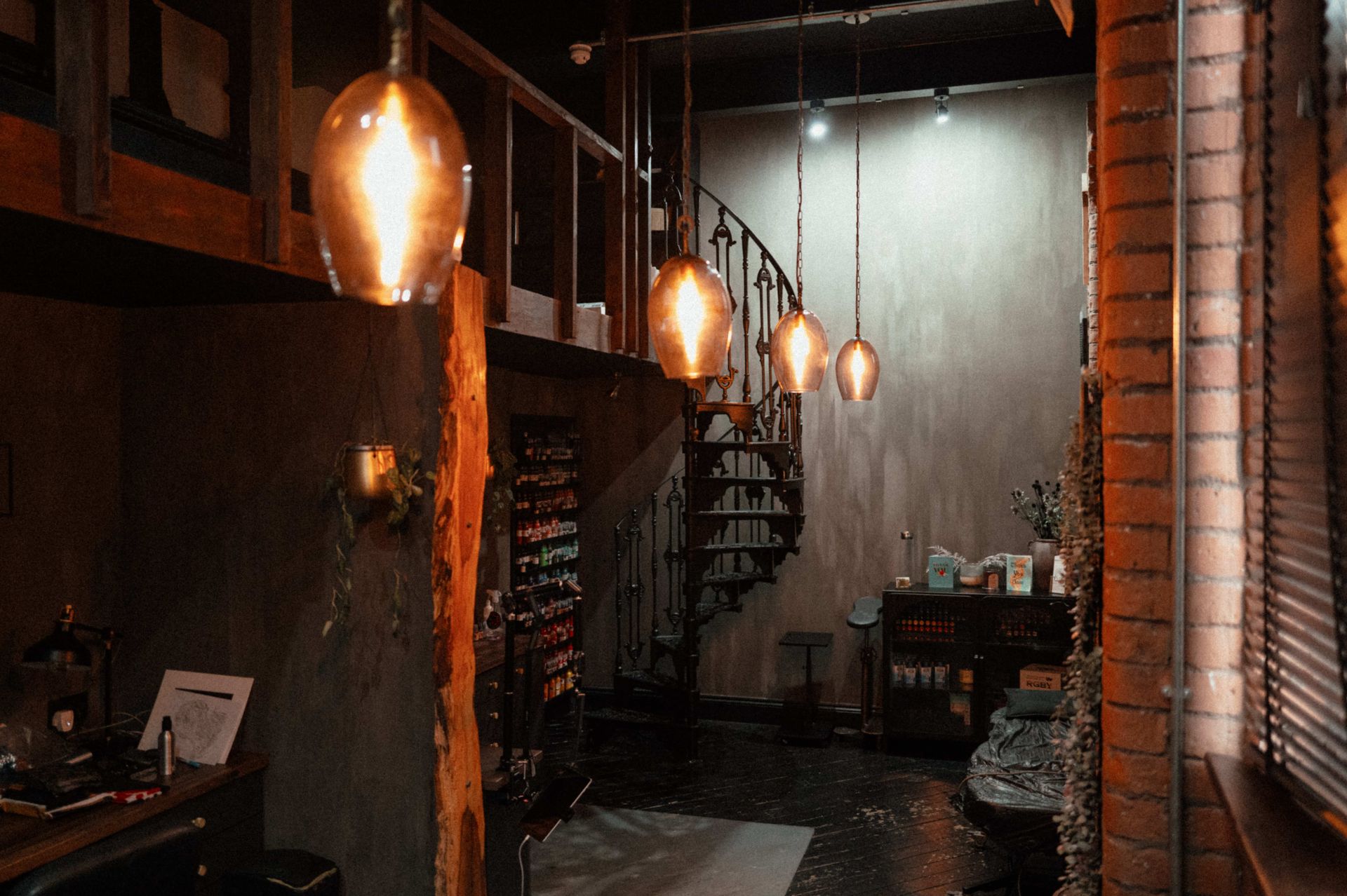 A dimly lit interior space with amber pendant lights, a spiral staircase, exposed brick, and a wooden loft structure.