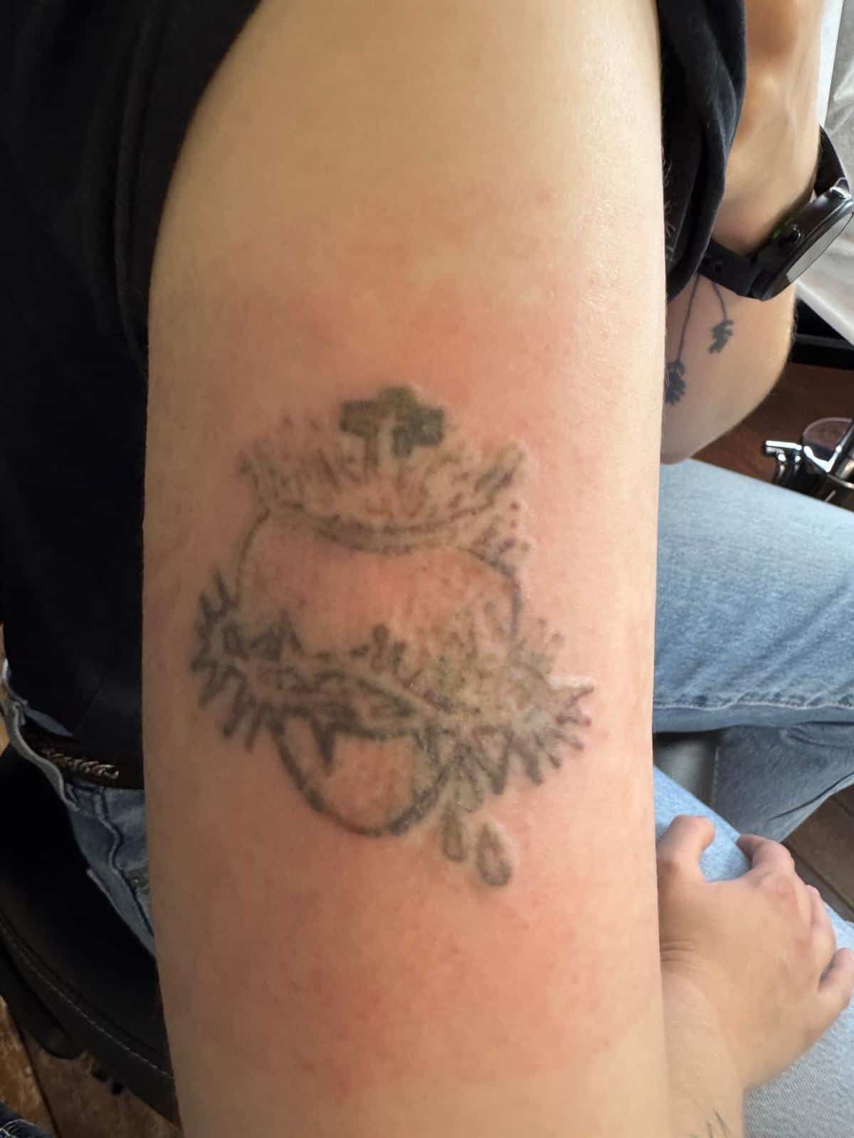A person’s upper arm featuring a black-ink tattoo of the Sacred Heart of Jesus surrounded by a crown of thorns and a cross.