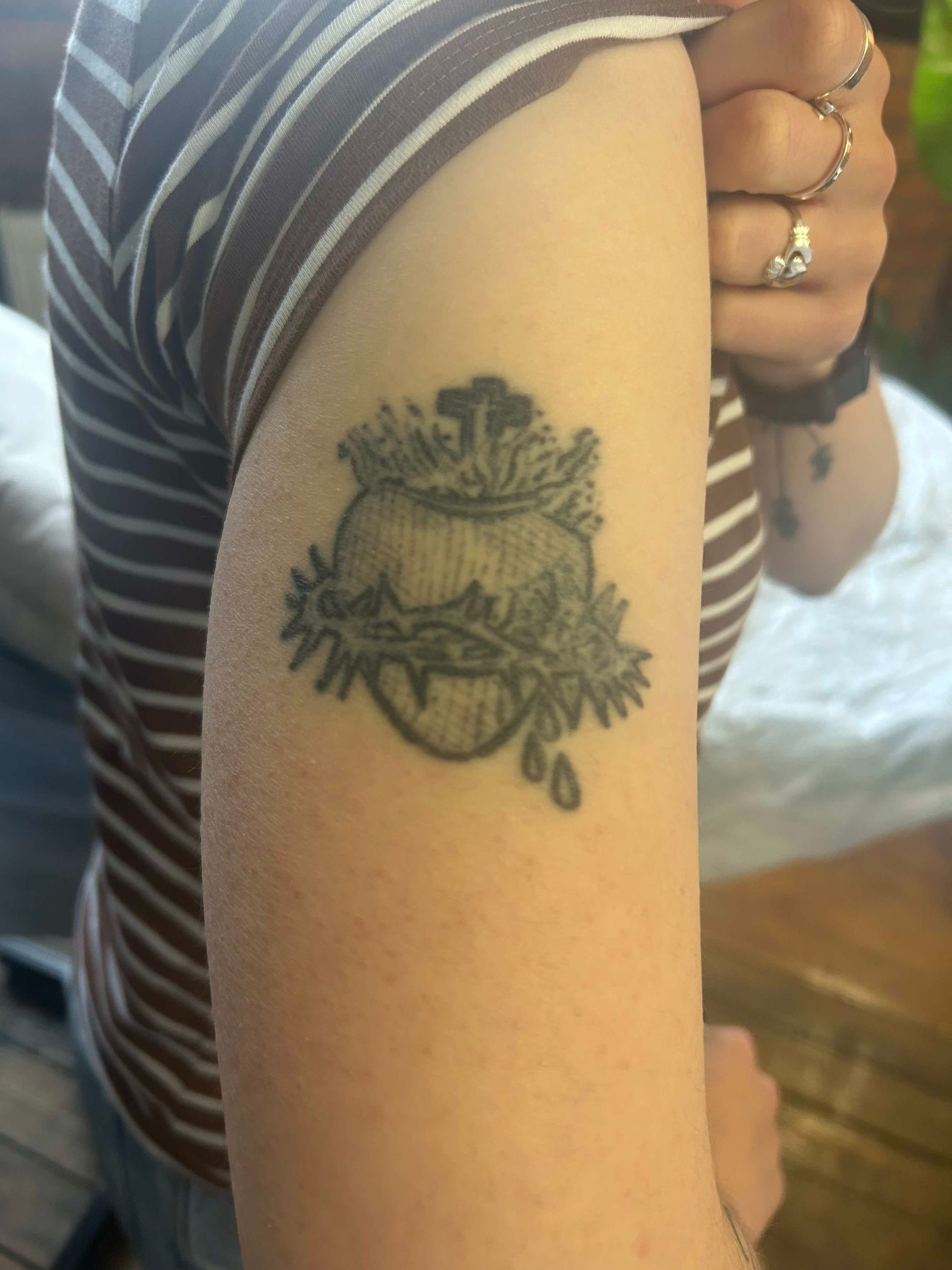 A close-up of a person's upper arm featuring a black ink tattoo of a Sacred Heart encircled by a crown of thorns.