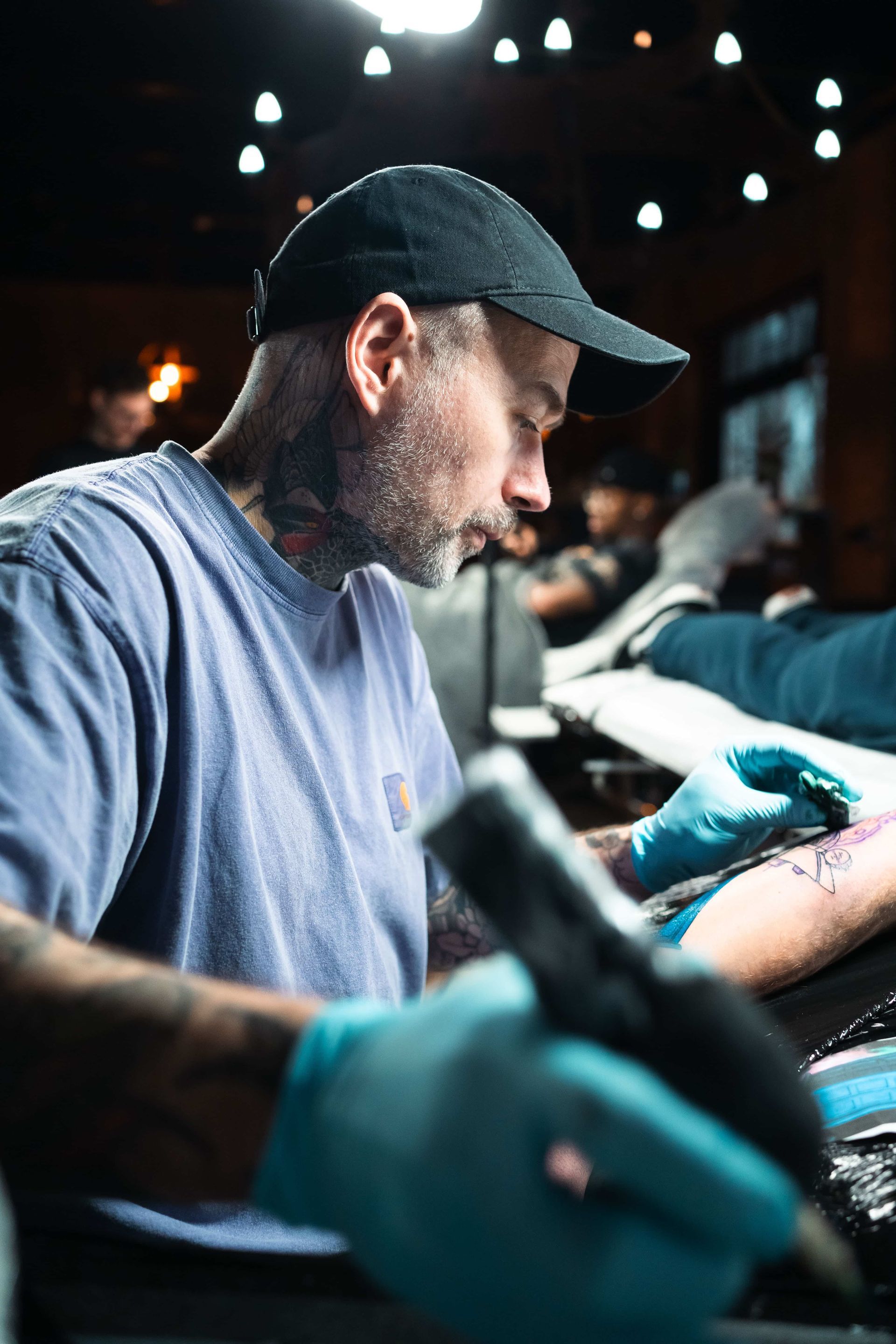 A tattoo artist wearing a black baseball cap and blue gloves focuses on tattooing a client in a dimly lit studio.