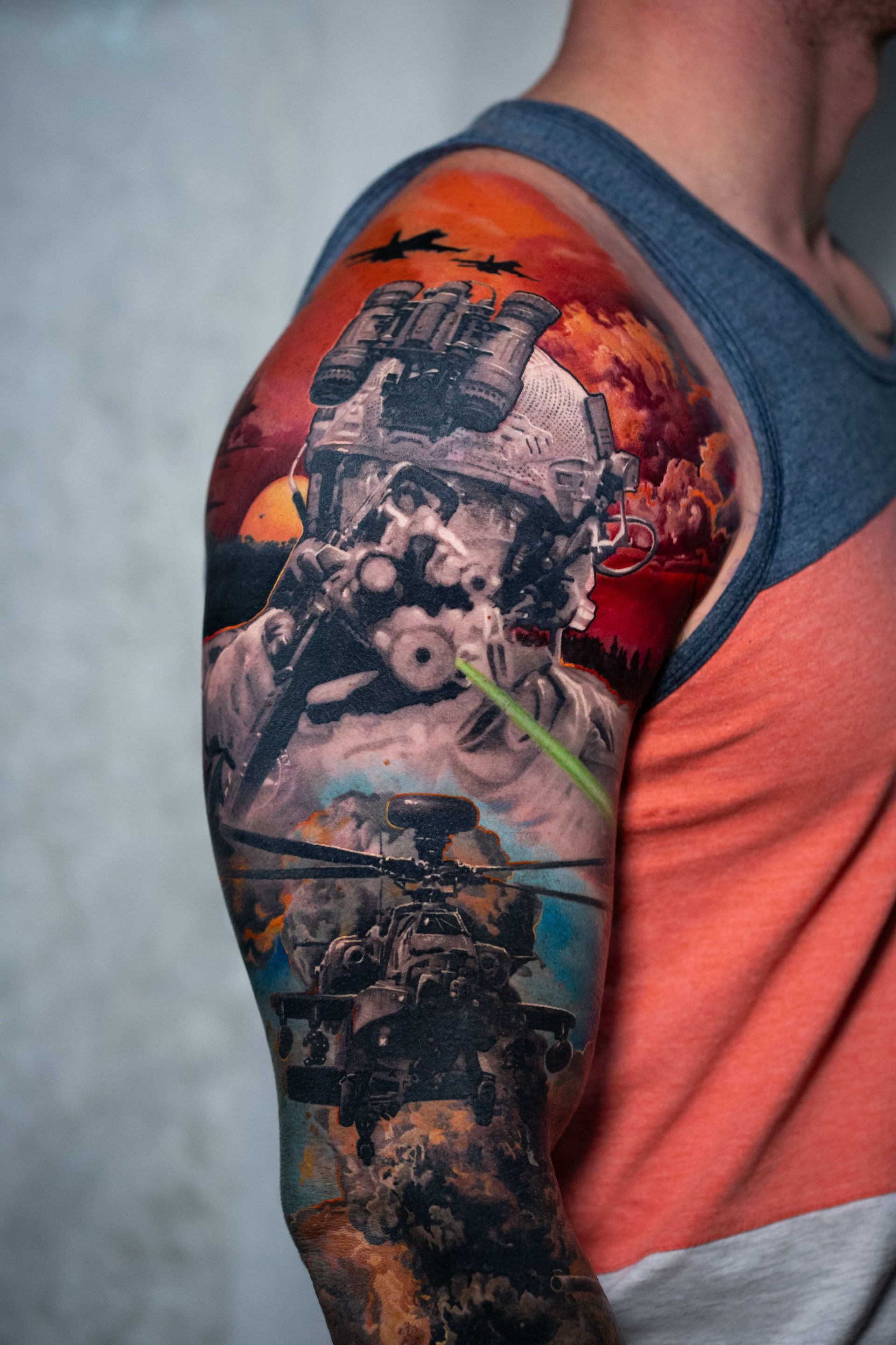 A shoulder tattoo featuring a soldier in tactical gear, a combat helicopter, and a dramatic orange, fiery sunset sky.