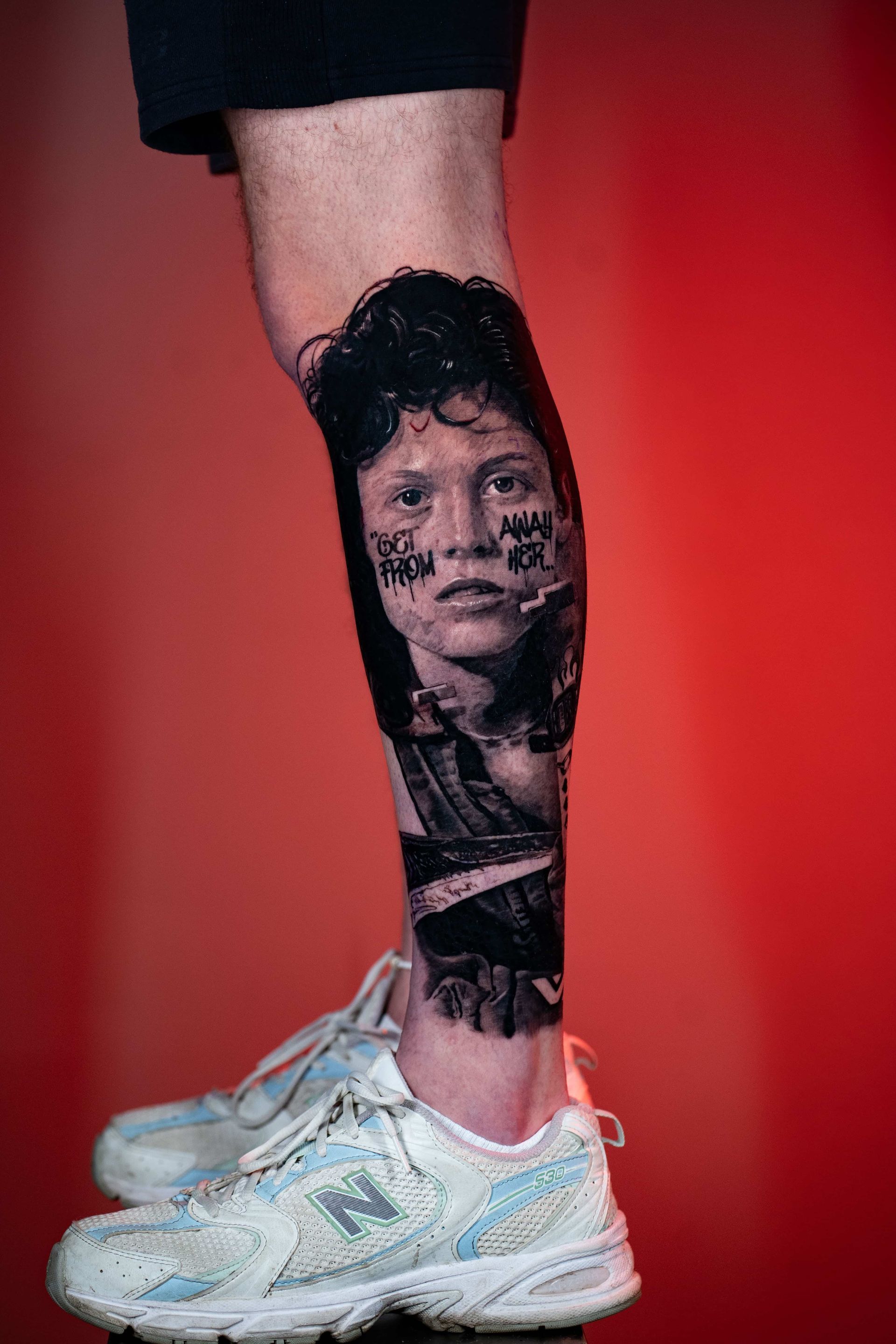 A detailed tattoo of a person with dark hair on a lower leg against a vibrant red background.