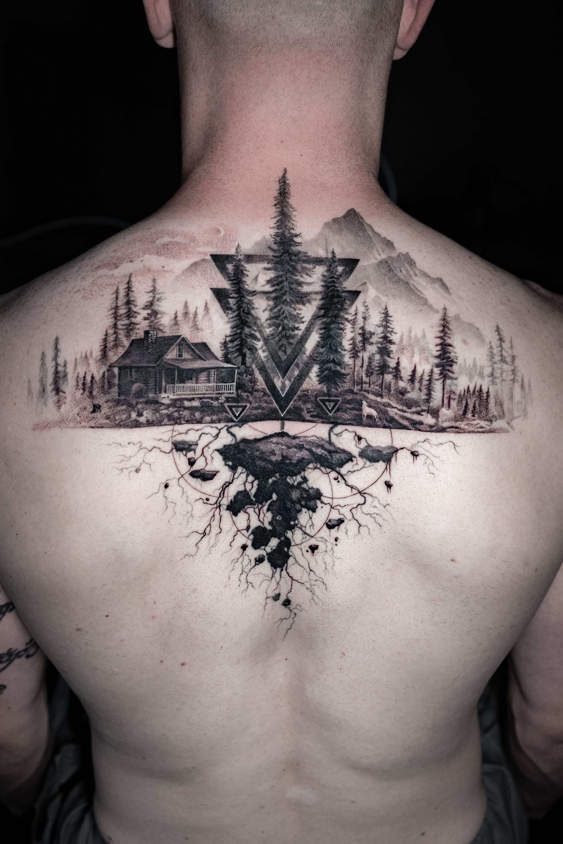 A black and grey tattoo on a person's upper back depicting a forest, cabin, mountains, and roots in a geometric style.