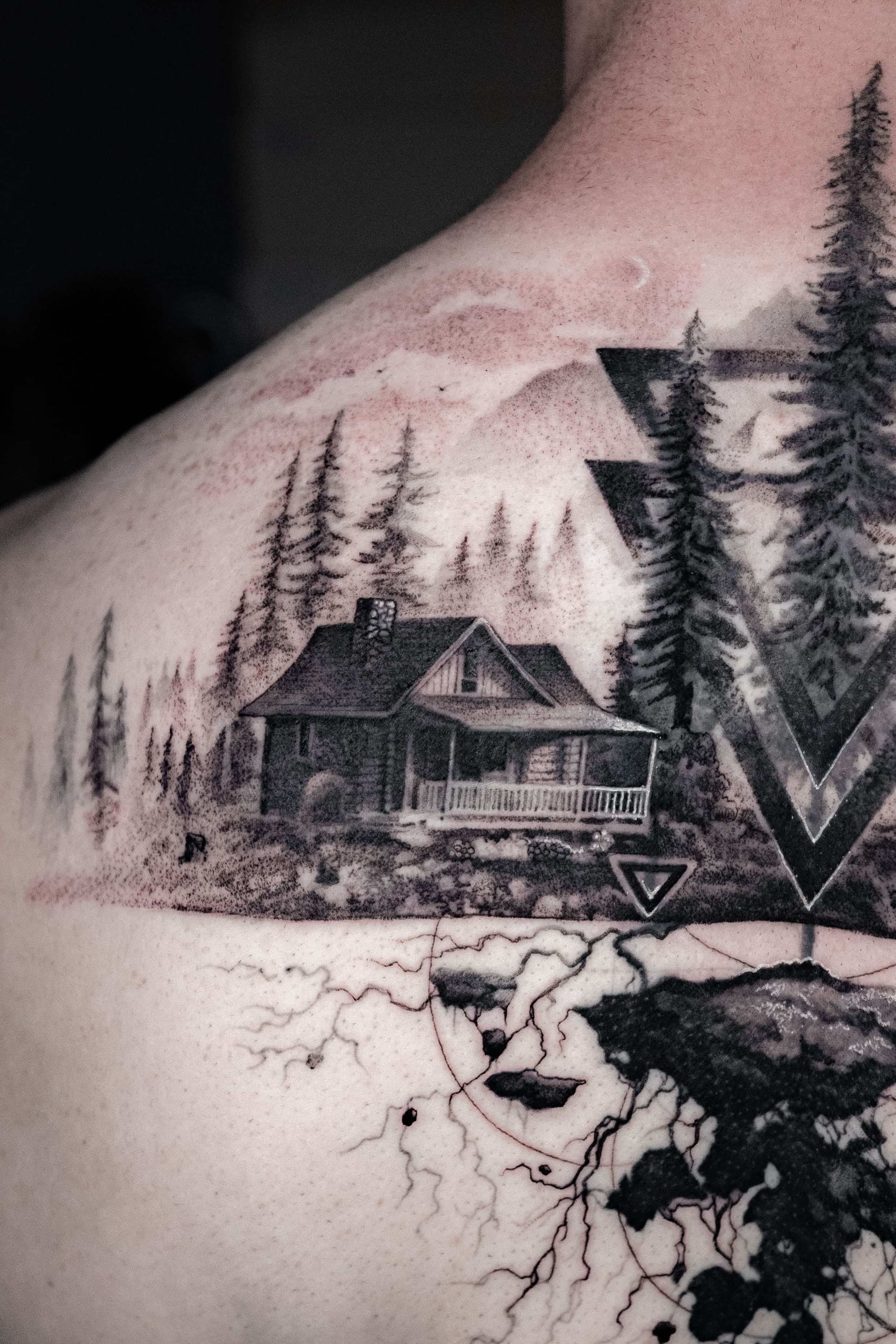 Black and grey tattoo on a shoulder featuring a rustic cabin in a forest, geometric triangles, and tree roots below.