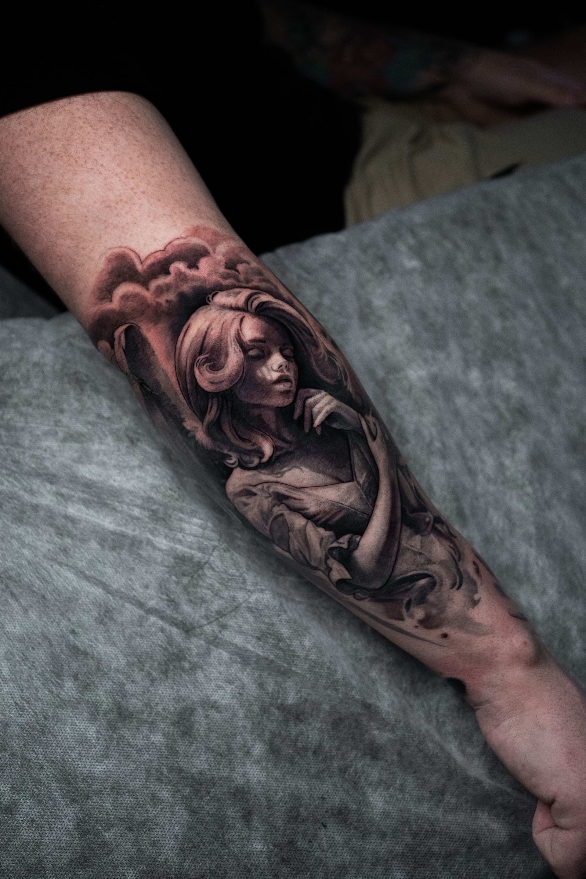 Black and gray realism tattoo on a forearm depicting a somber, winged figure emerging from shadowy clouds.