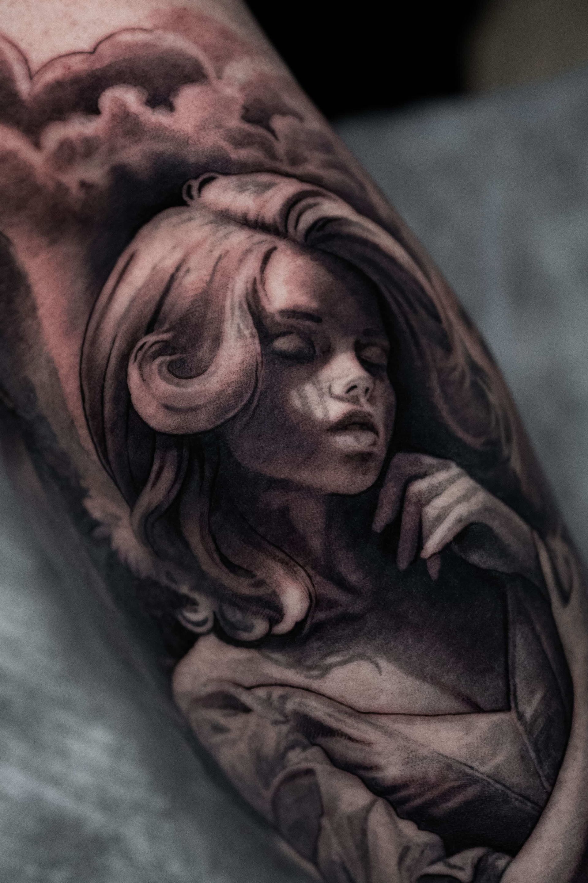 A black-and-grey tattoo of a person with wavy hair, eyes closed, and hand near their face, framed by flowing clouds.