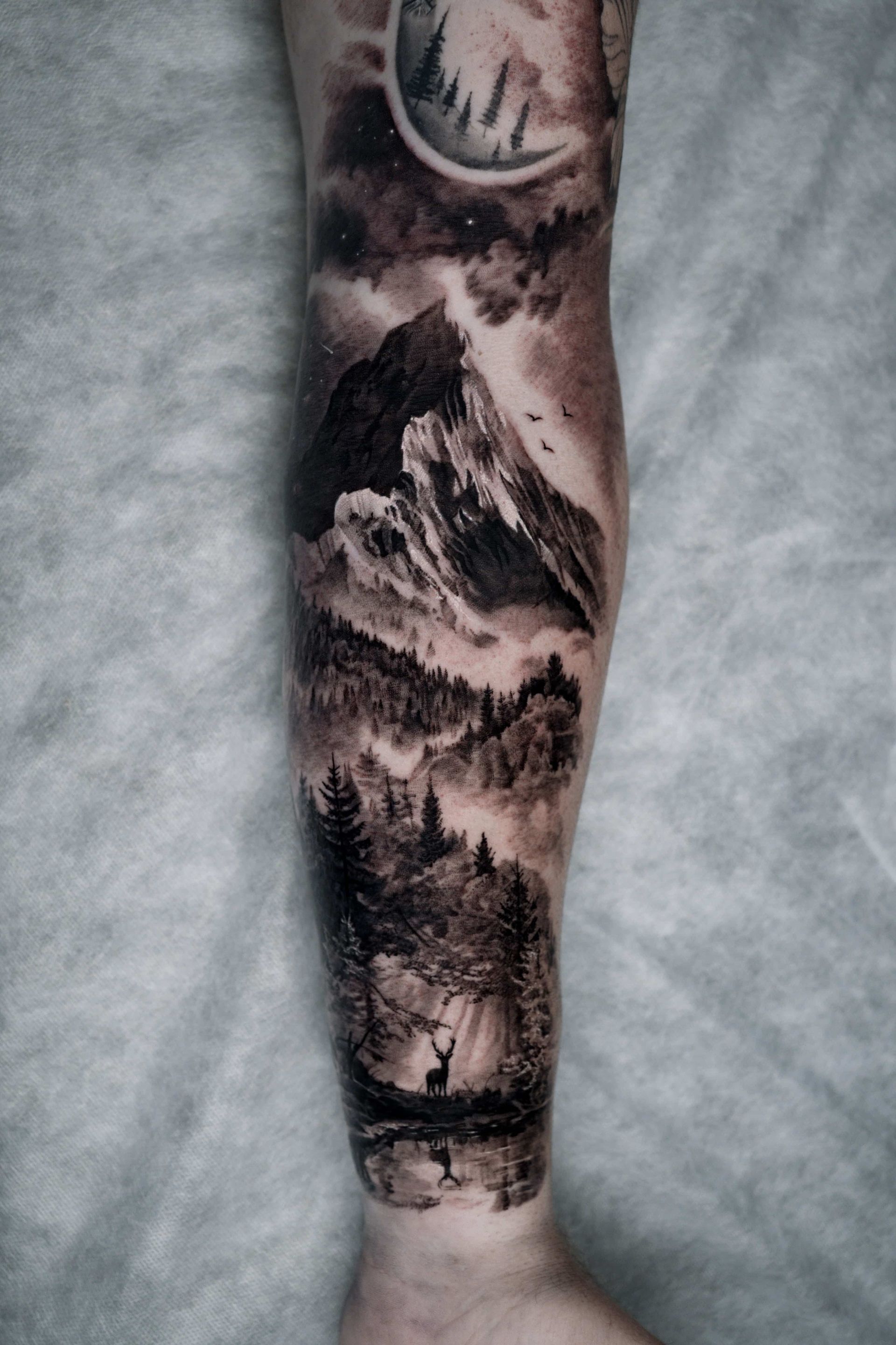 Black and grey tattoo on a lower leg featuring a crescent moon above mountain peaks, a forest, and a deer by a lake.