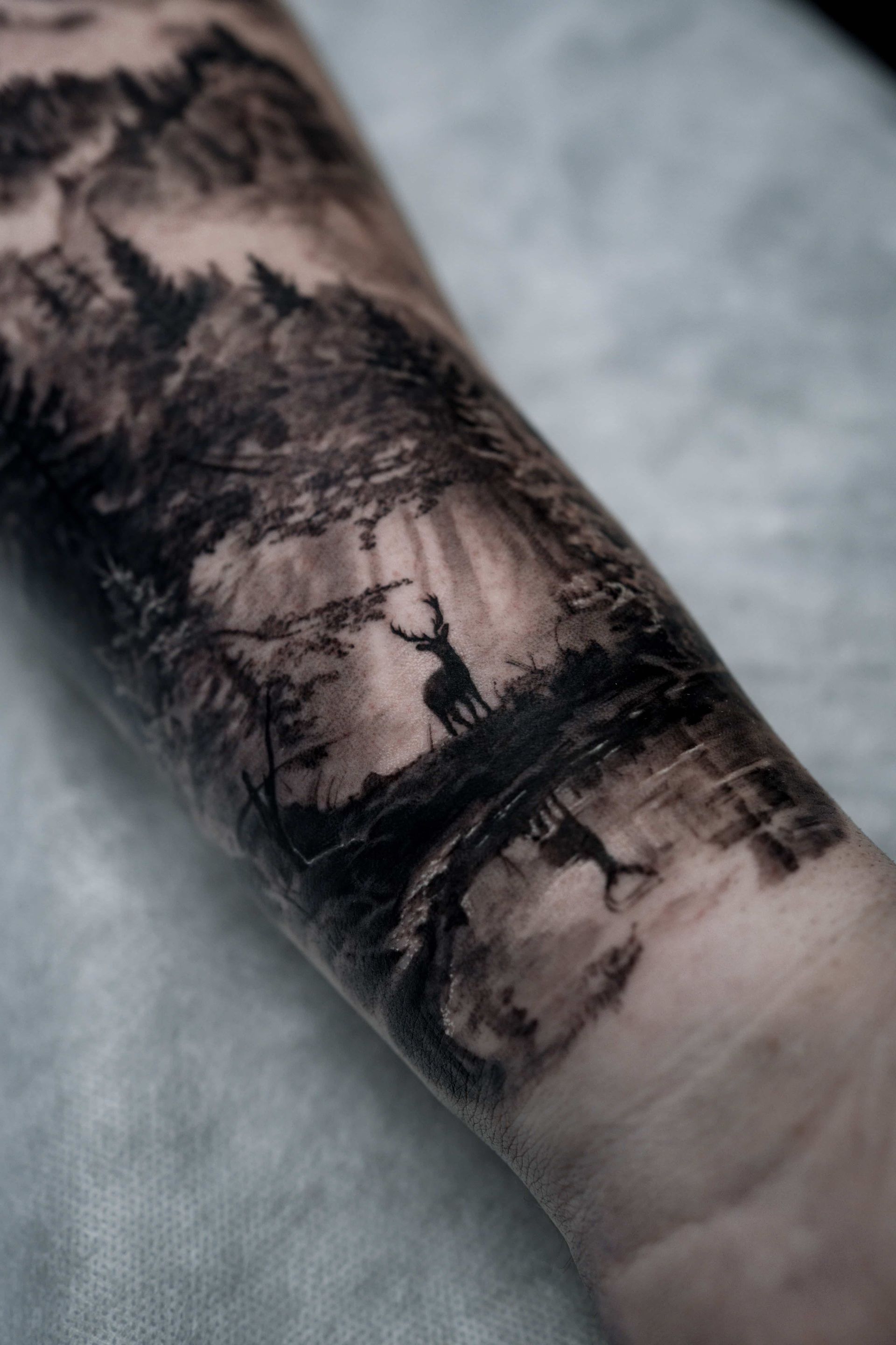 A detailed black-and-grey tattoo on an arm showing a deer standing in a forest beside a reflective body of water.