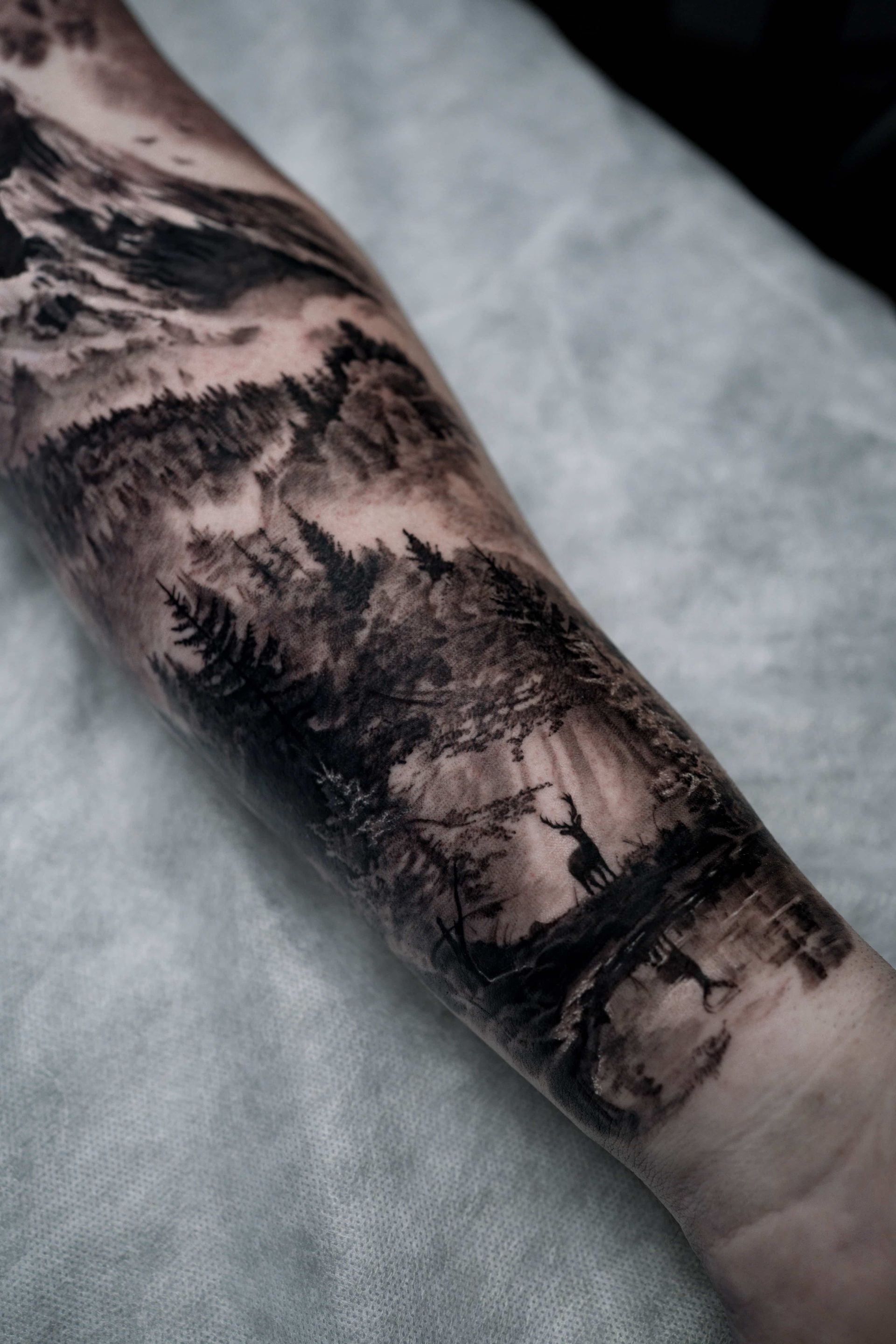 Black and gray forearm tattoo featuring a detailed mountain range, dense evergreen forest, and a deer by a lake.