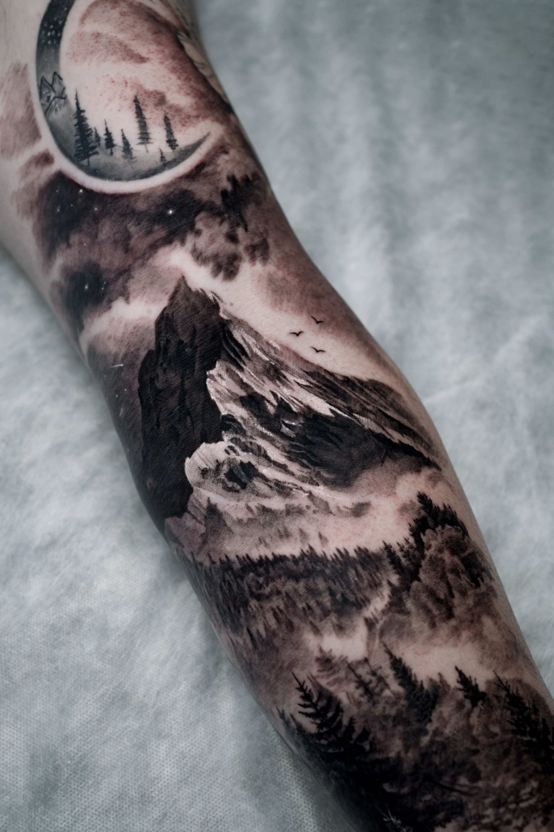 Black and gray tattoo of a mountain range, pine forest, and a crescent moon with landscape detail against a starry sky.