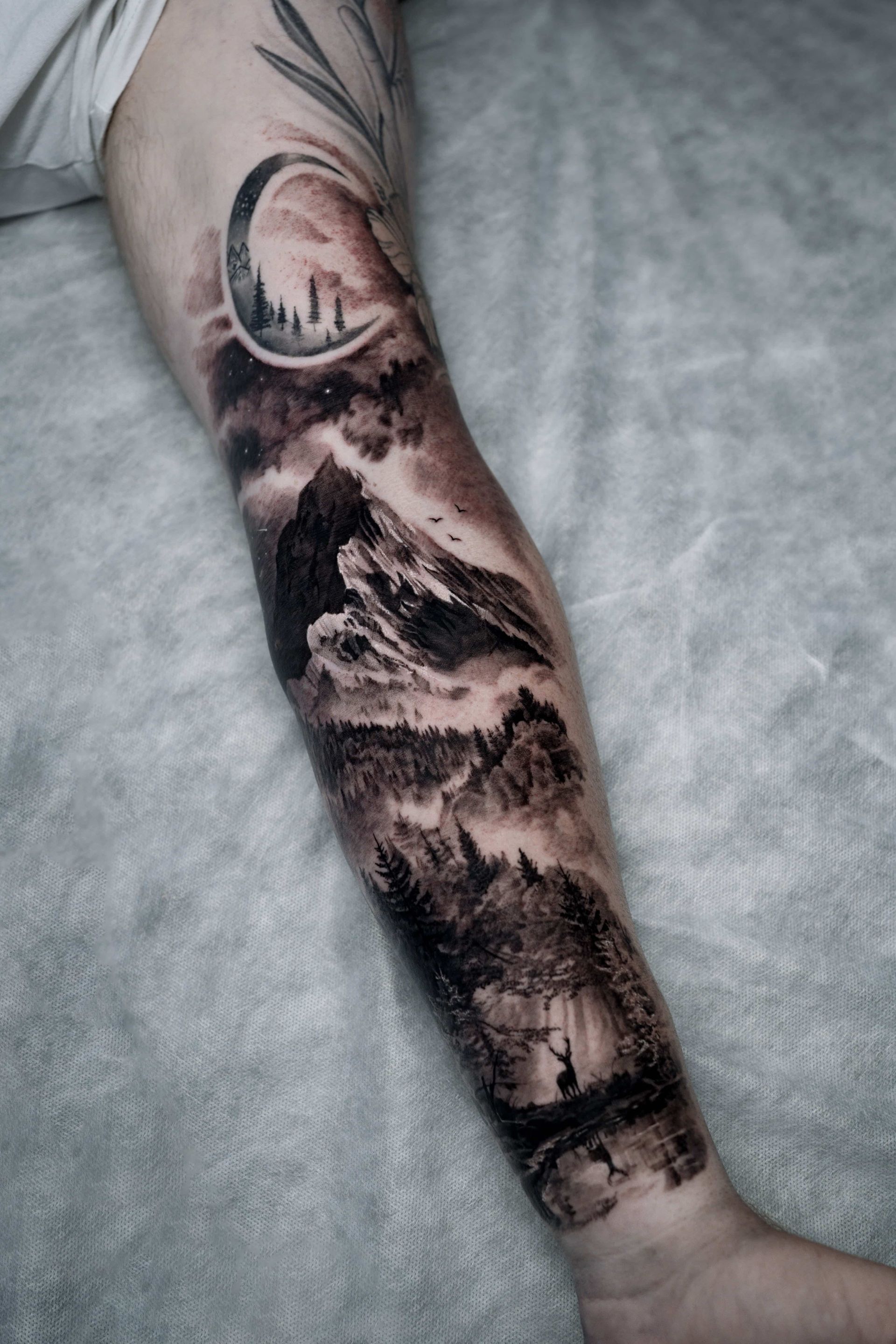 A black-and-grey tattoo on an arm featuring a mountain landscape, pine forest, a crescent moon, and a deer by a stream.