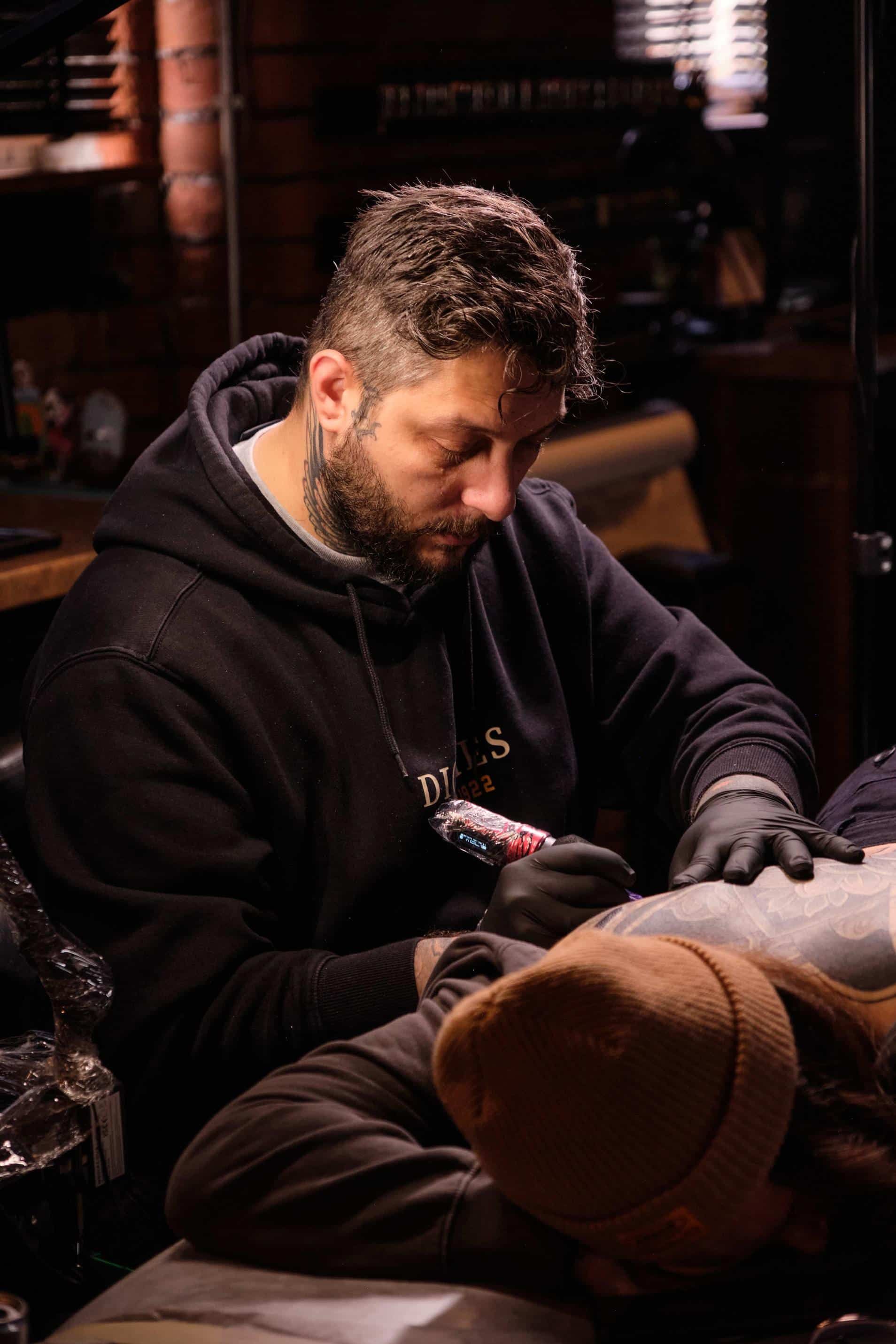 A focused tattoo artist in a black hoodie wearing gloves tattoos a client in a dim, wood-paneled studio.