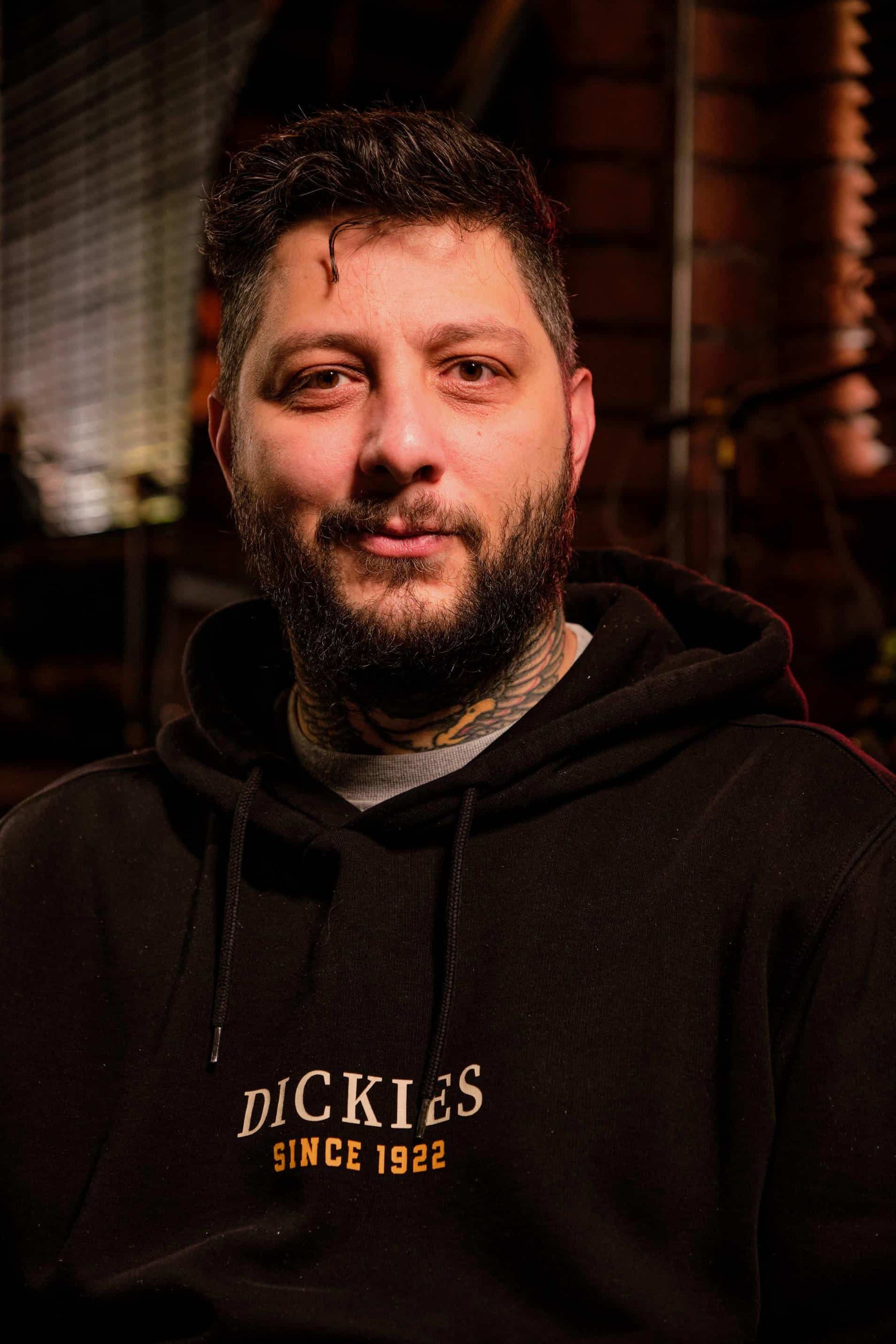 A person with a beard and tattoos on their neck wears a black Dickies hooded sweatshirt, looking directly at the camera.