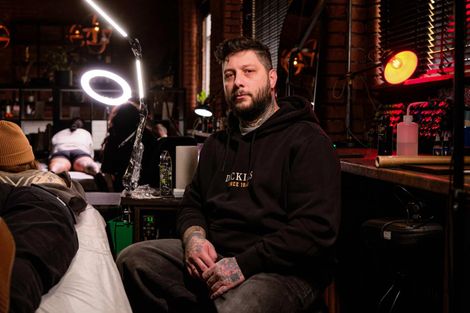 A tattooed artist sits in a dimly lit, industrial-style tattoo studio, with equipment and clients in the background.
