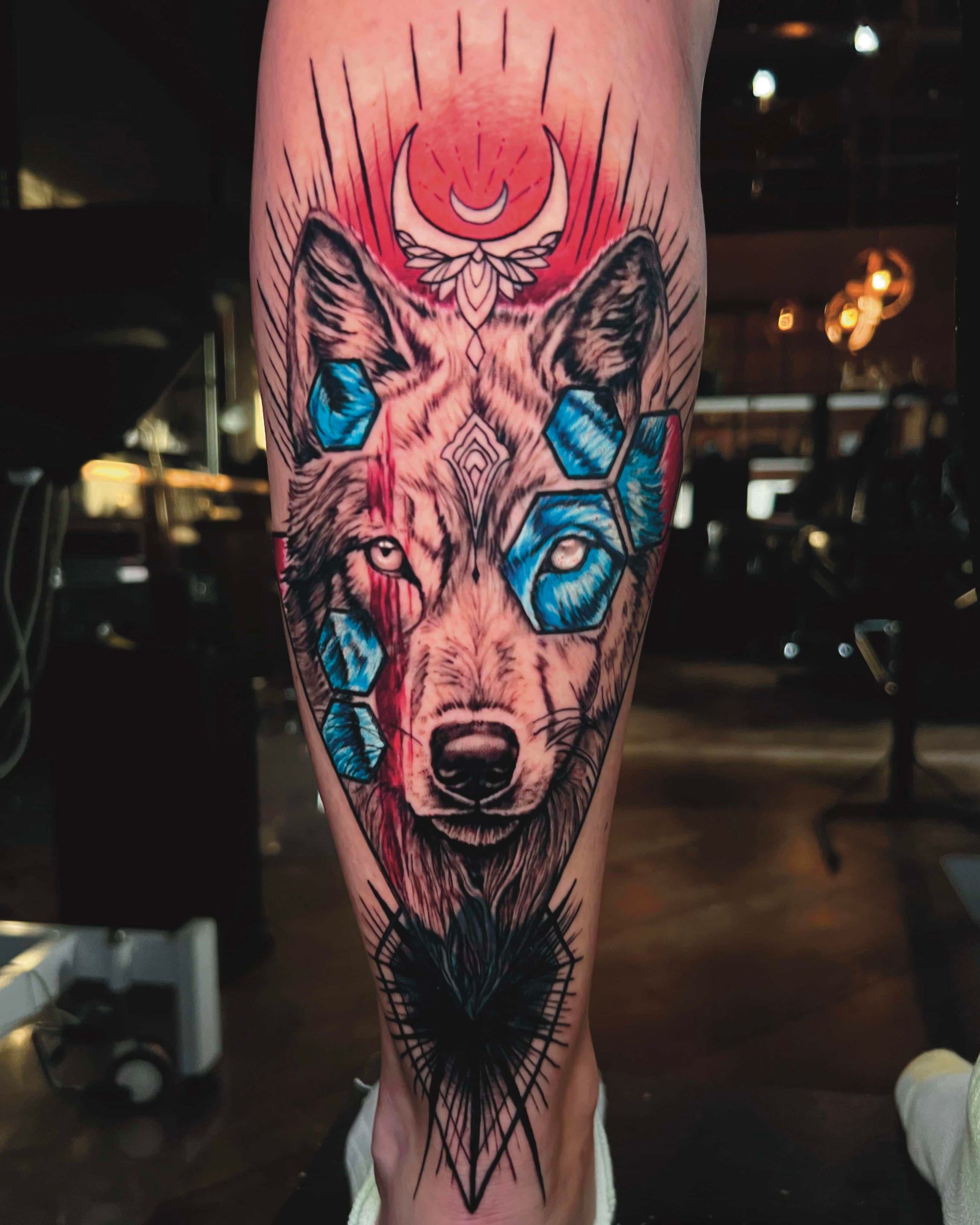 A detailed tattoo of a wolf's face featuring blue geometric shapes, a red sunburst background, and a crescent moon.