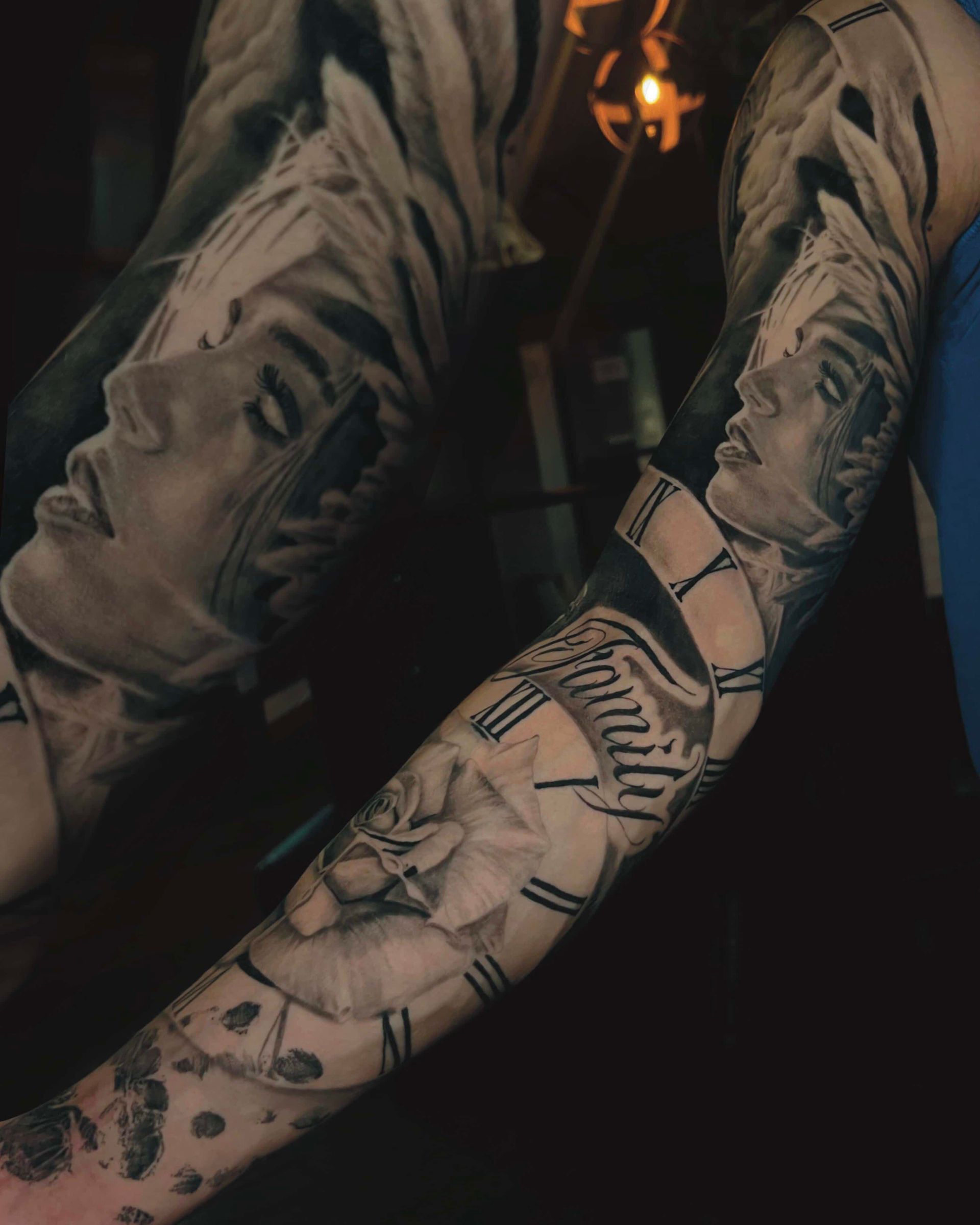 Close-up of a detailed black-and-grey sleeve tattoo featuring a face, rose, clock, and the word