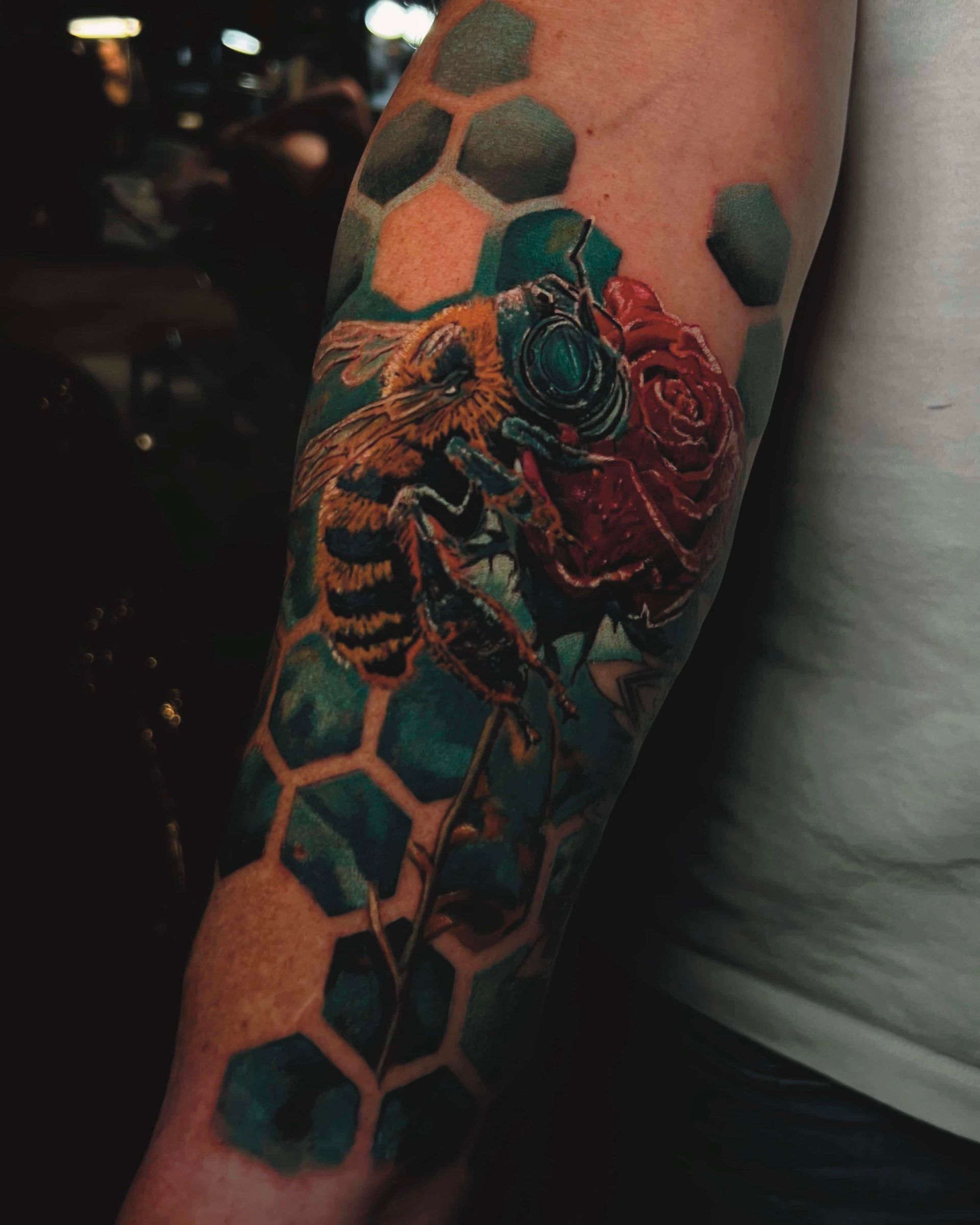 A forearm tattoo featuring a detailed bee on a red rose, layered over a dark, hexagonal honeycomb background pattern.