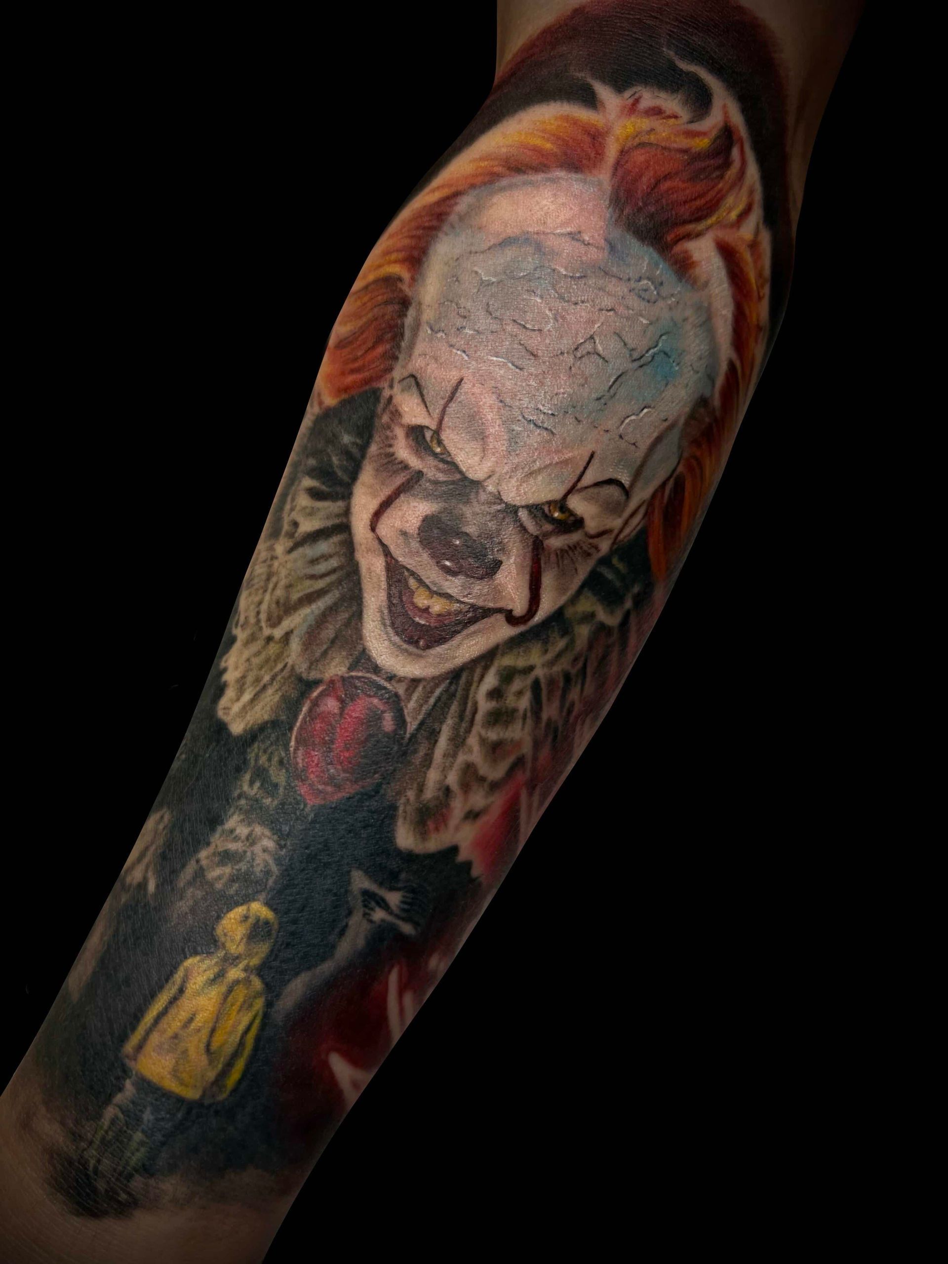 A forearm tattoo of Pennywise the Clown looking down at a small figure in a yellow raincoat against a dark background.
