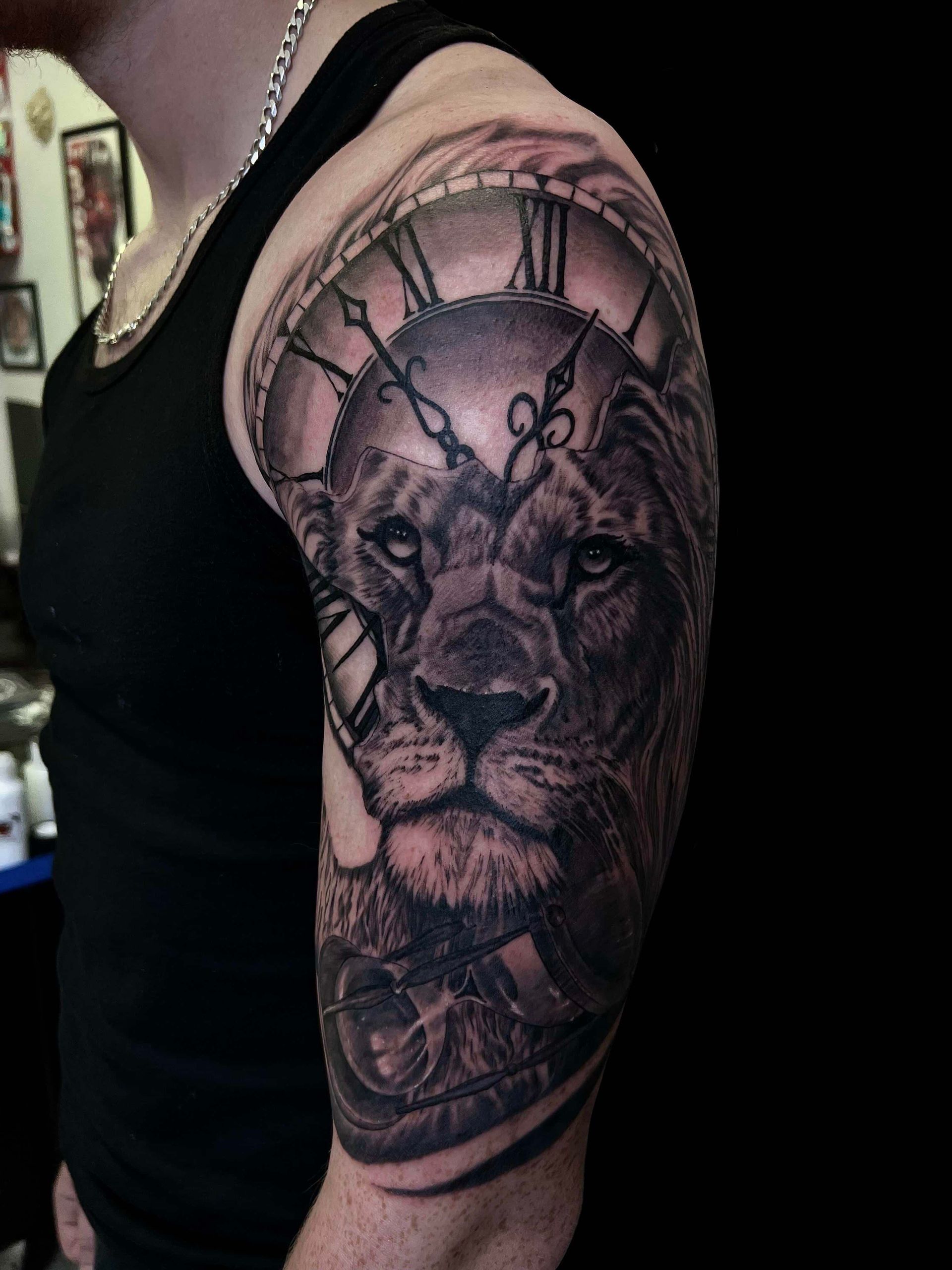A realistic black-and-grey tattoo on an upper arm featuring a lion’s face partially framed by a large clock face.