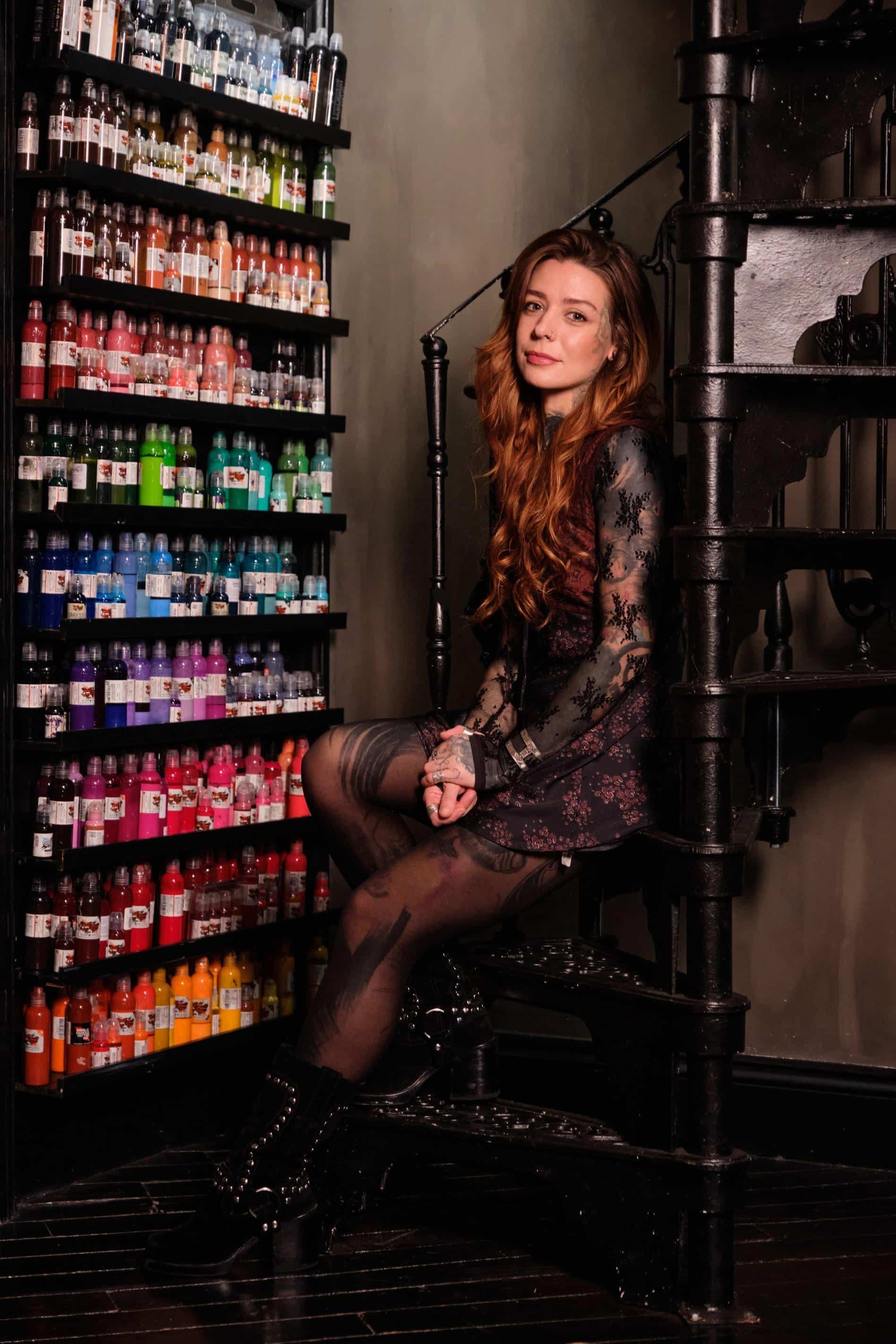 A person with long reddish hair and tattoos sits on a spiral staircase beside a wall filled with shelves of ink bottles.