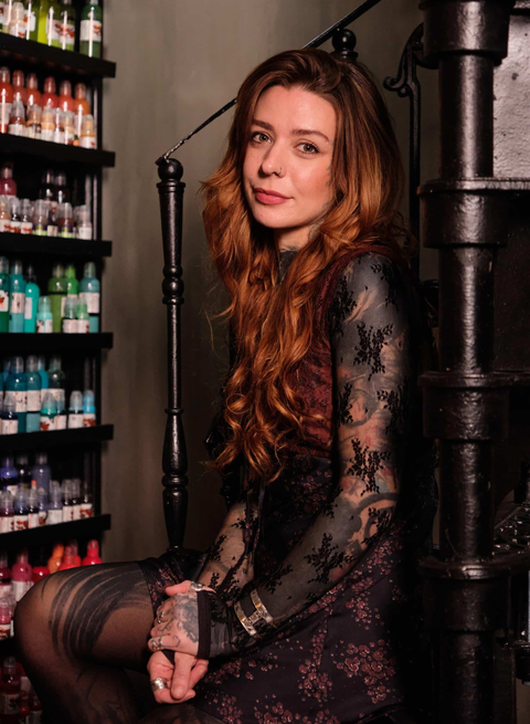 A person with long, wavy auburn hair wearing a lace-sleeved dress, sitting on a staircase beside shelves of colorful bottles.