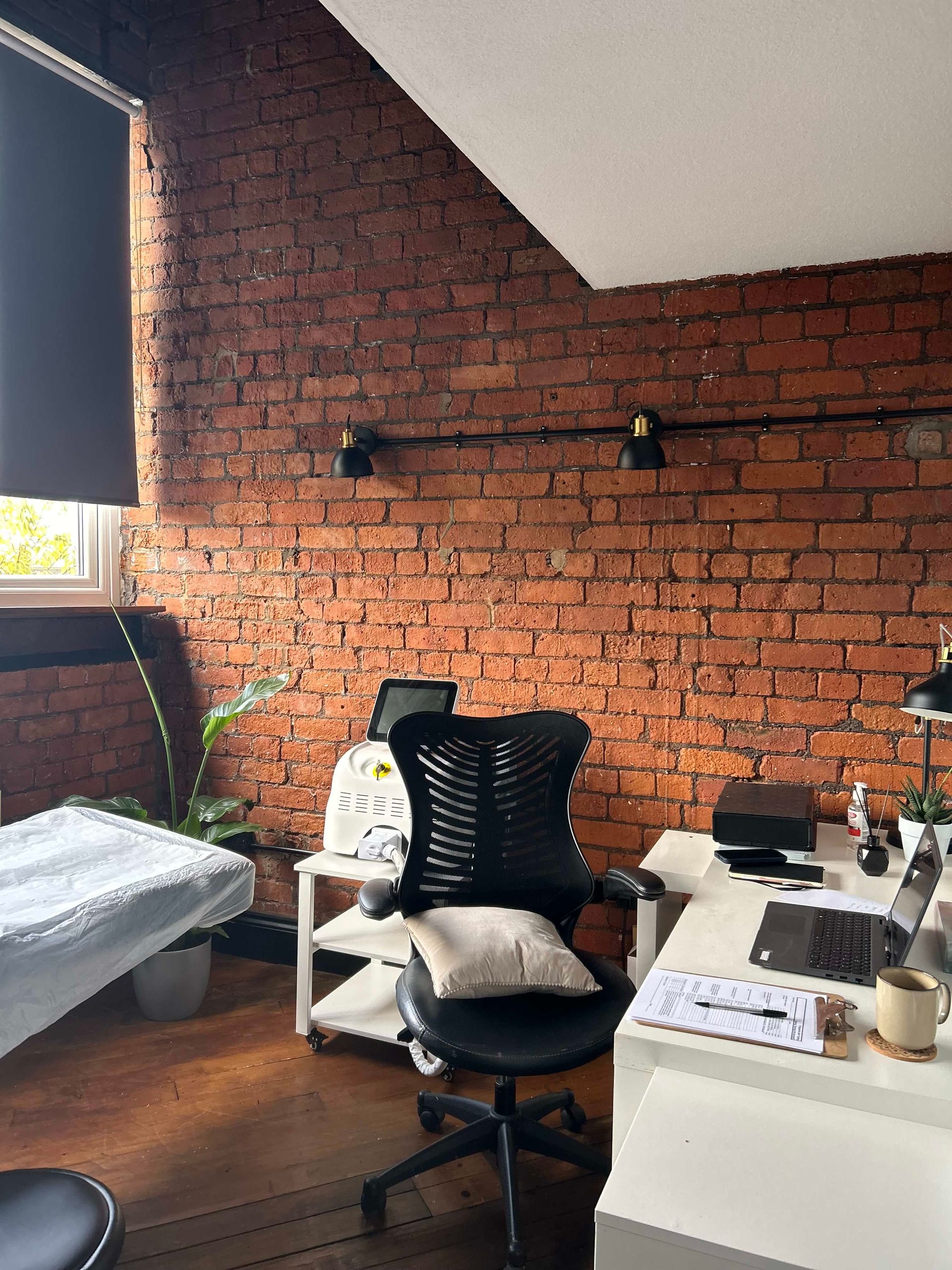 A cozy home office with a brick wall, an ergonomic chair, a white desk with a laptop, and a houseplant in a corner.