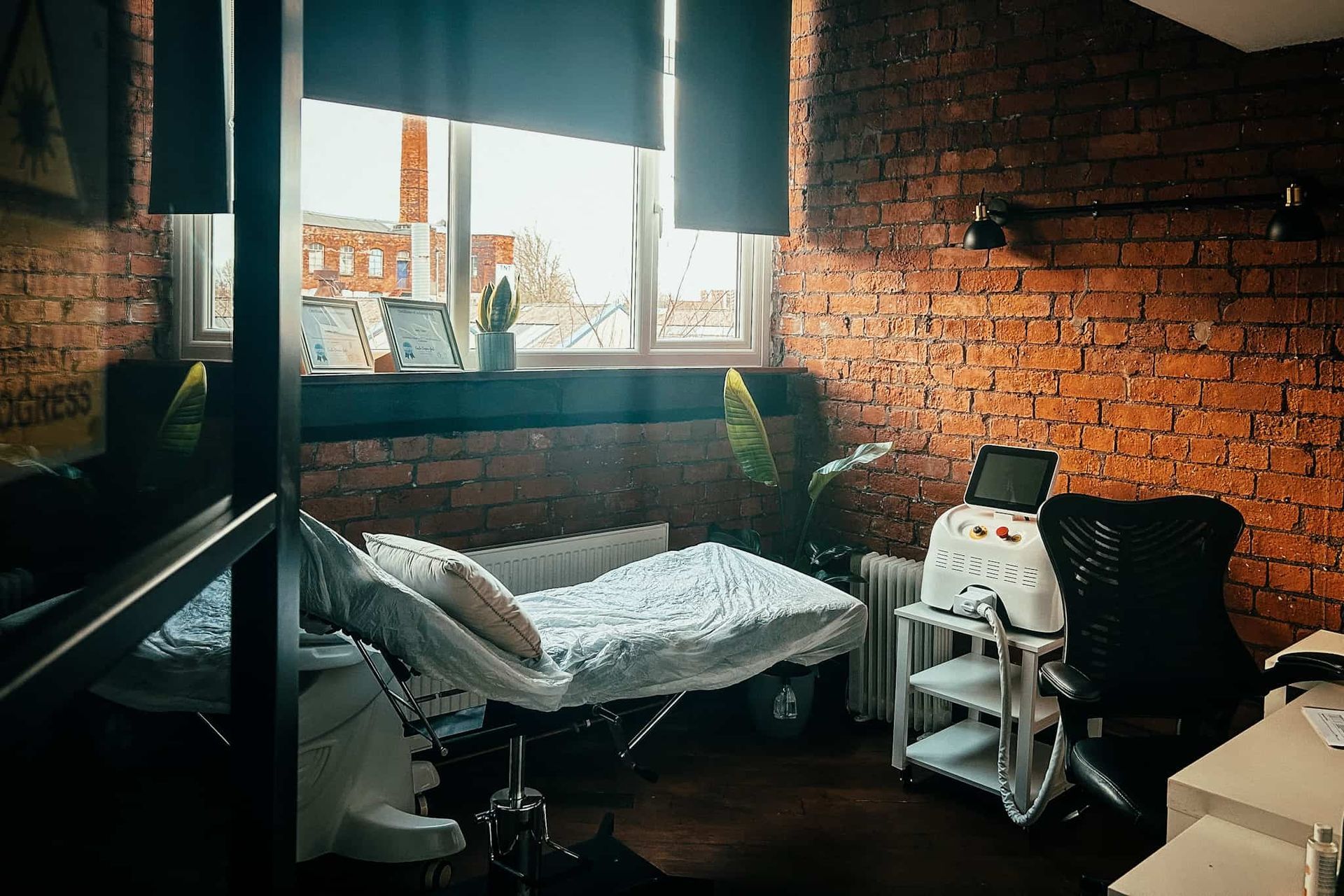 A treatment room featuring an examination bed, a specialized medical device on a rolling cart, and a chair near a window.