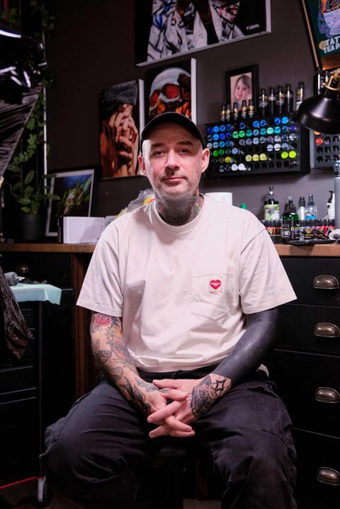 Tattoo artist with neck and arm tattoos sitting in a studio with supplies and artwork in the background.