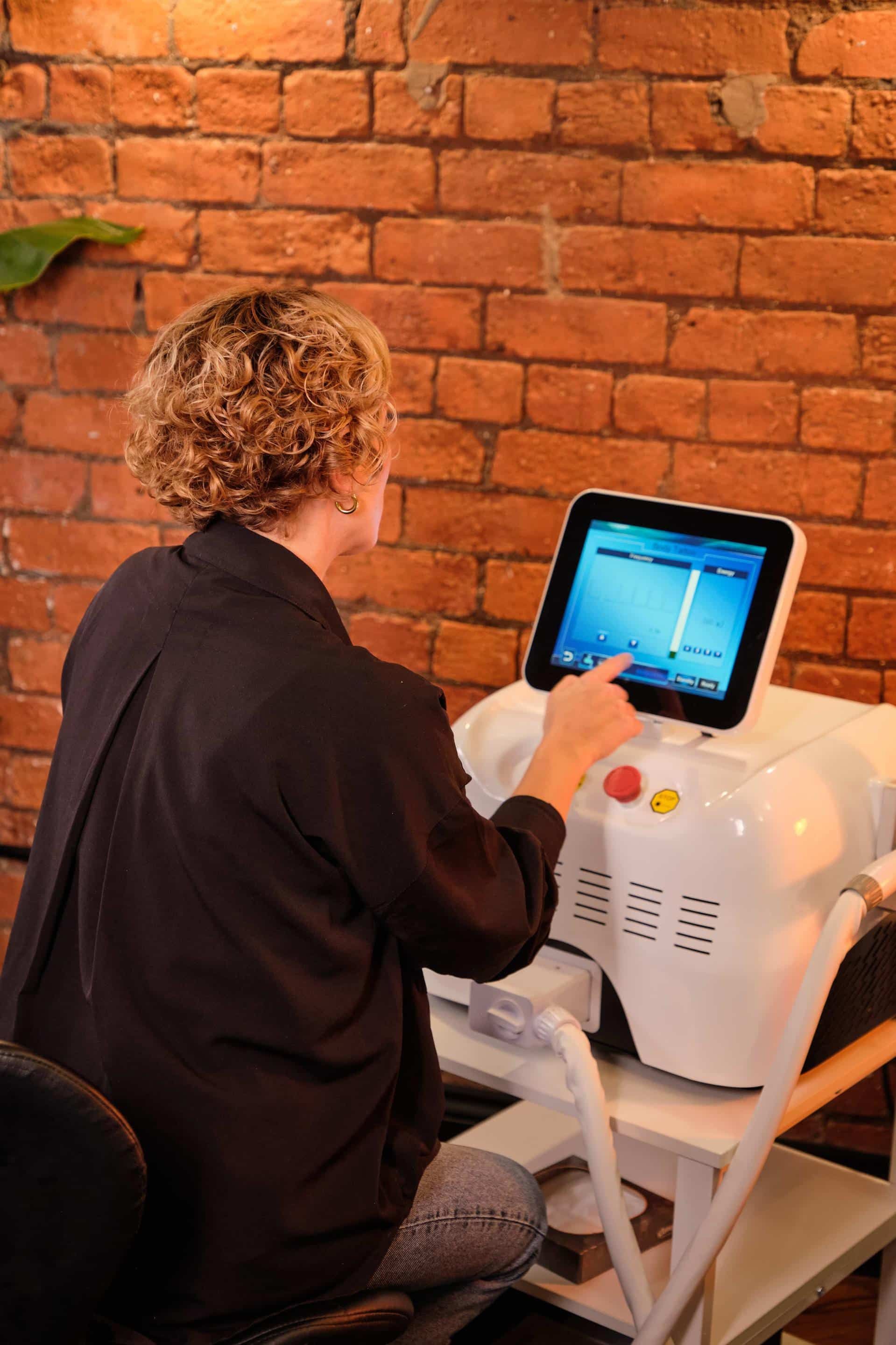 A person with curly hair adjusts settings on a professional medical touchscreen device against a brick wall background.