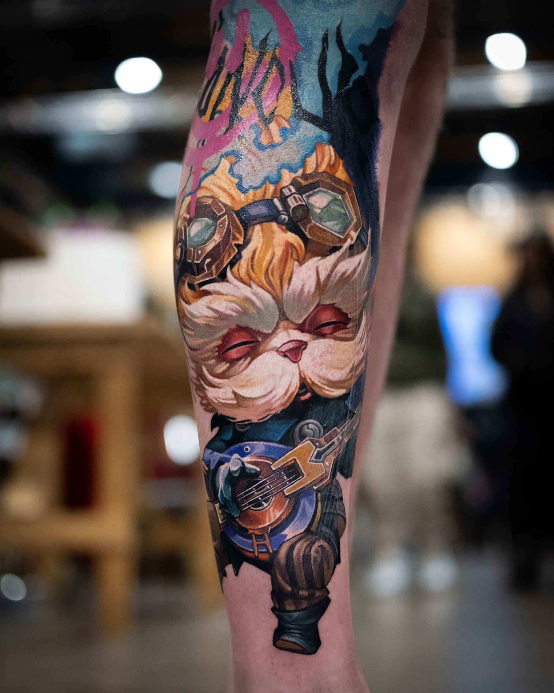 A colorful tattoo on a leg featuring a stylized, fuzzy character with goggles playing a small, wooden guitar.