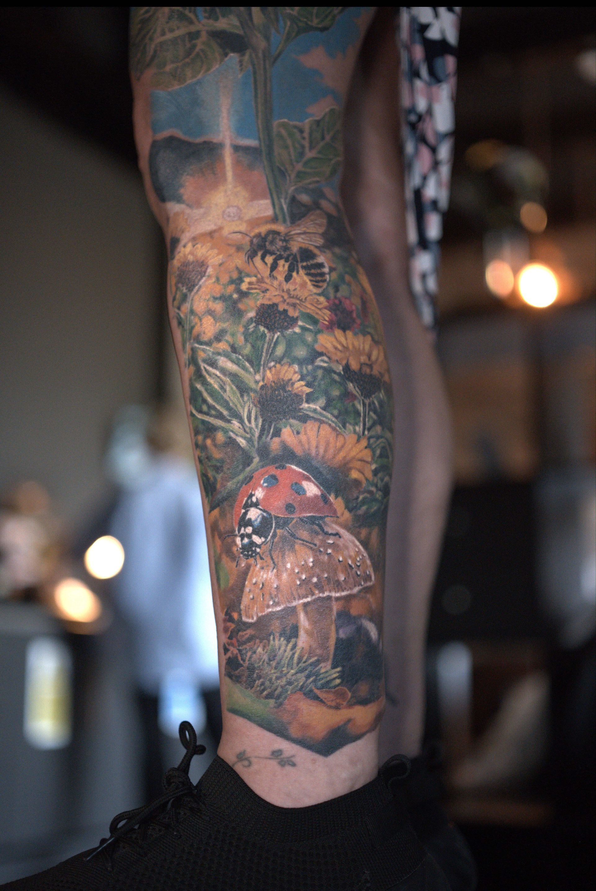 A detailed color tattoo on a lower leg featuring a mushroom, a ladybug, and various wildflowers in a forest setting.