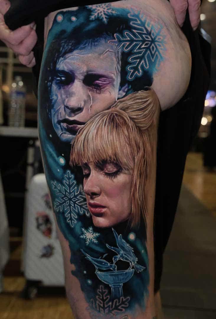 A detailed color tattoo on a thigh featuring two realistic faces surrounded by blue snowflakes and abstract shapes.