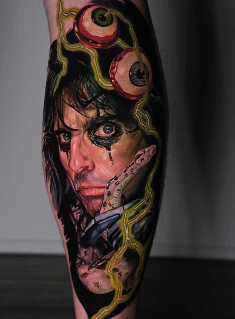 A vibrant, detailed tattoo of Alice Cooper with dark eye makeup, surrounded by floating eyeballs and a snake on a dark bg.