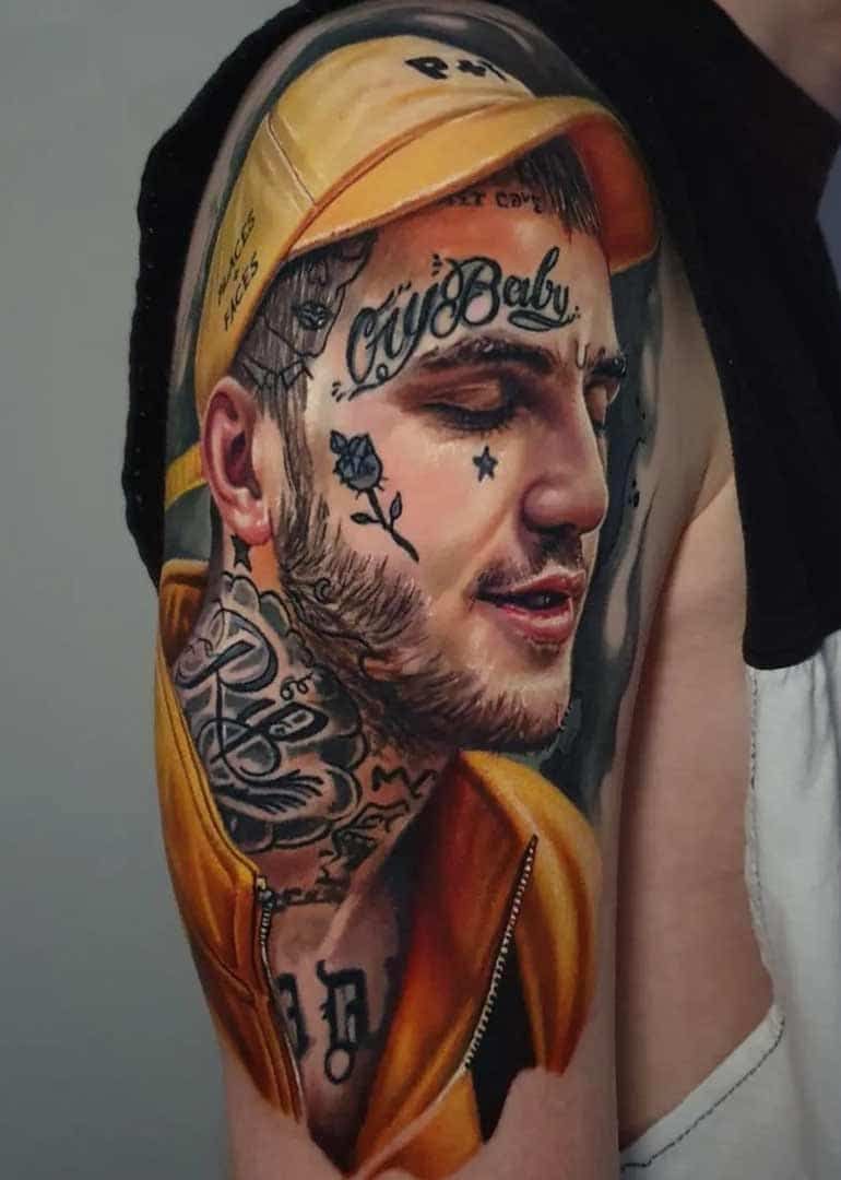 A realistic portrait tattoo of Lil Peep on a person's upper arm, featuring his signature face tattoos and yellow hat.