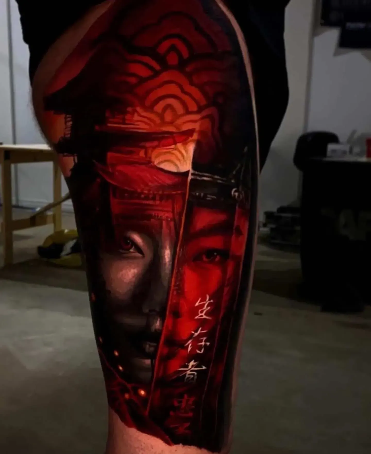 A vibrant red, surreal tattoo on a leg featuring a split face, a gate-like structure, and stylized Asian characters.