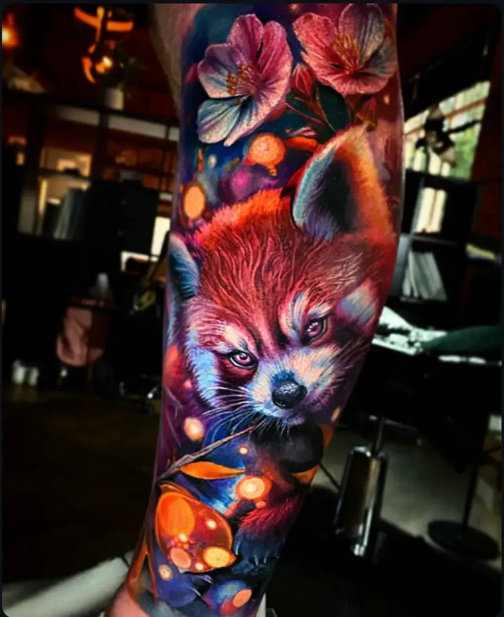 Vibrant, colorful tattoo of a red panda among flowers and glowing orange lights on someone's arm.