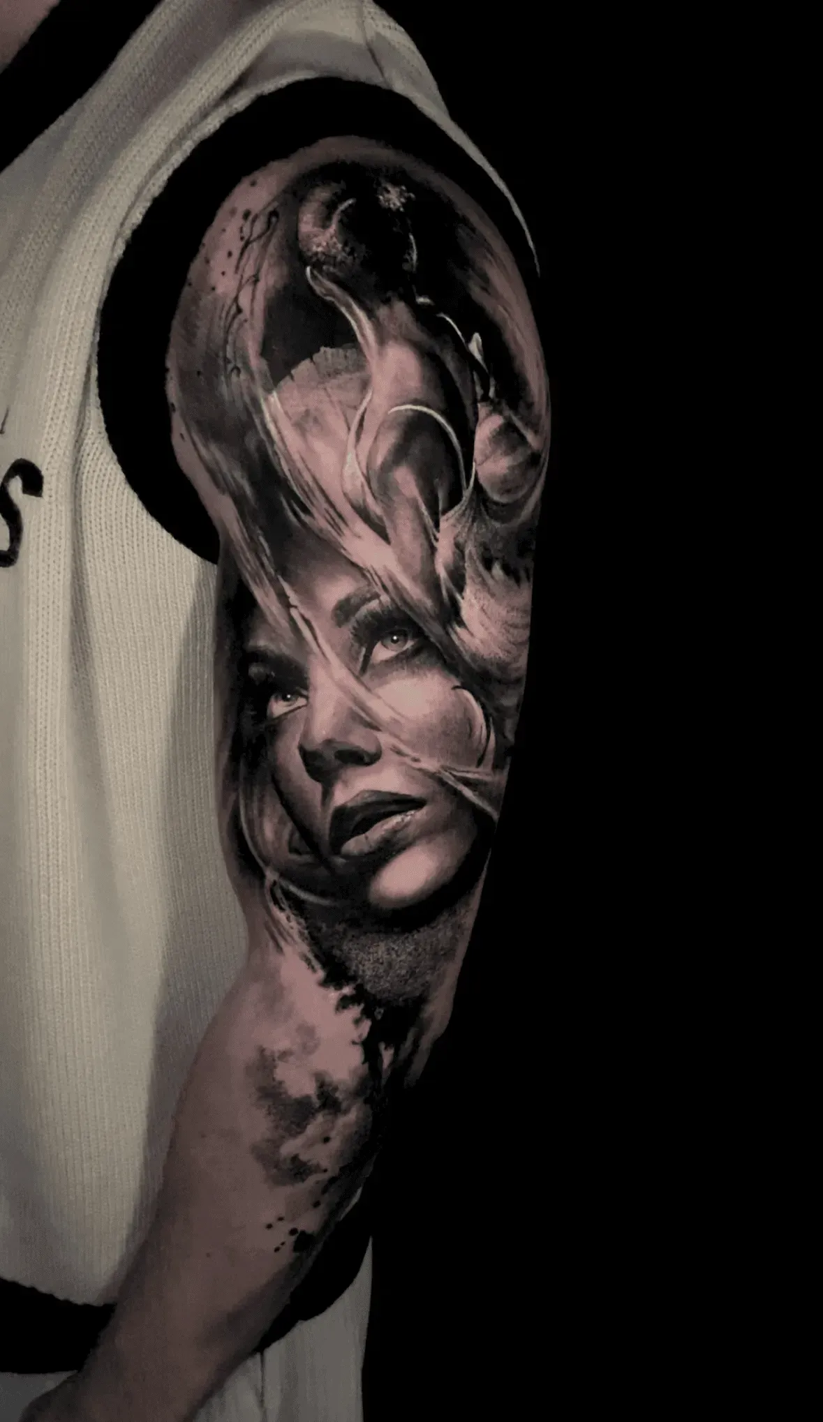 A realistic, monochromatic portrait tattoo on an upper arm featuring a face surrounded by flowing, smoky abstract patterns.