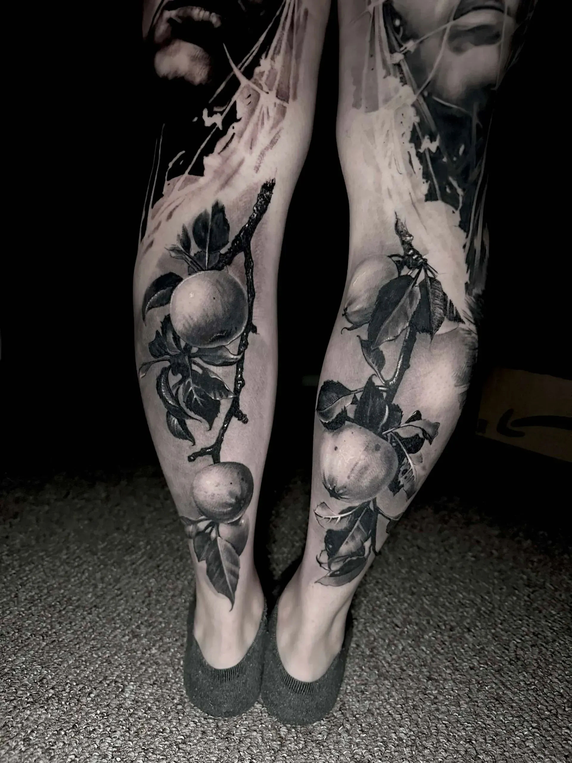 A pair of lower legs featuring realistic black and gray tattoos of fruit-bearing branches.