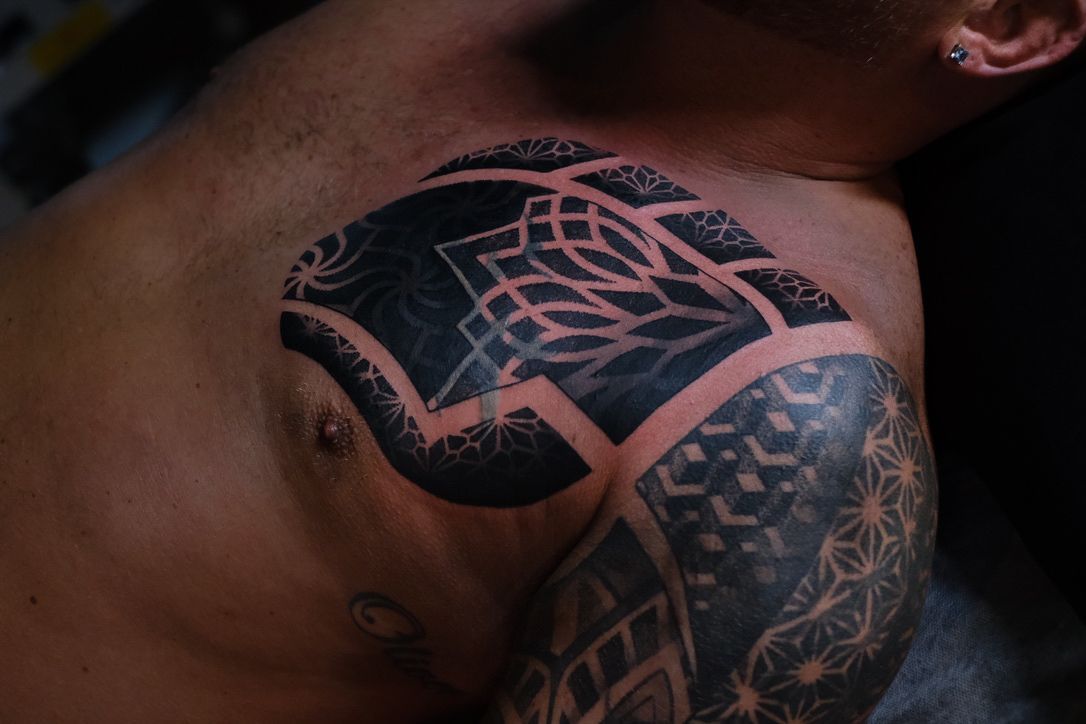 A person with blackwork geometric tattoos on their chest and shoulder, featuring intricate repeating patterns.