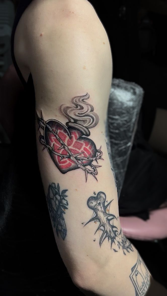 A person’s arm featuring multiple tattoos, highlighted by a red heart wrapped in barbed wire with rising wisps of smoke.