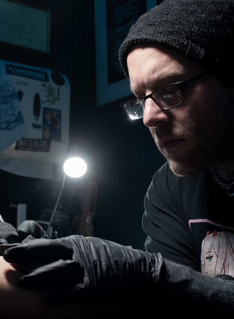 A person wearing a beanie, glasses, and black gloves works on a tattoo under a bright desk lamp in a dimly lit studio.