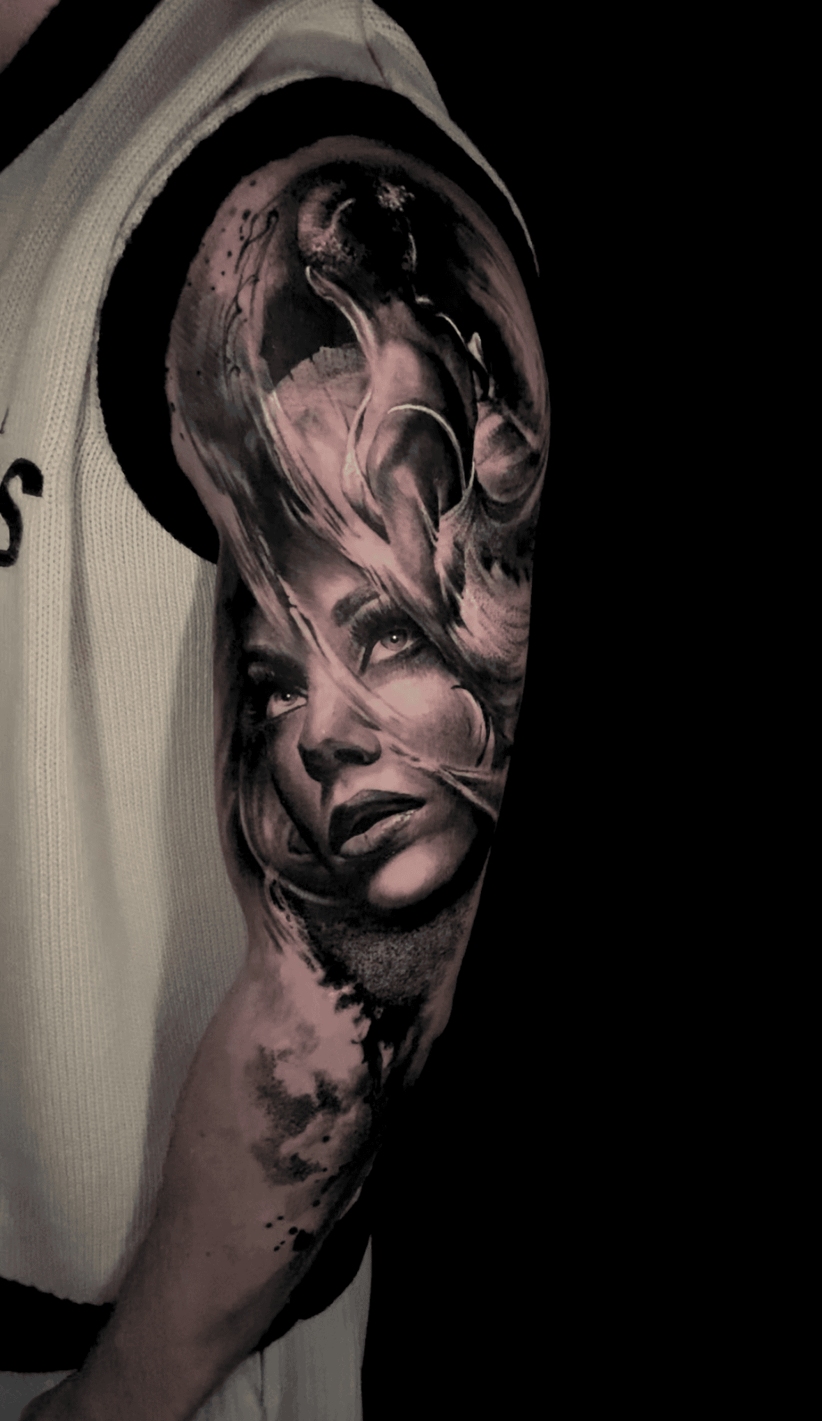 A realistic, black-and-grey tattoo on a person's upper arm featuring a face partially obscured by swirling, smoky shapes.