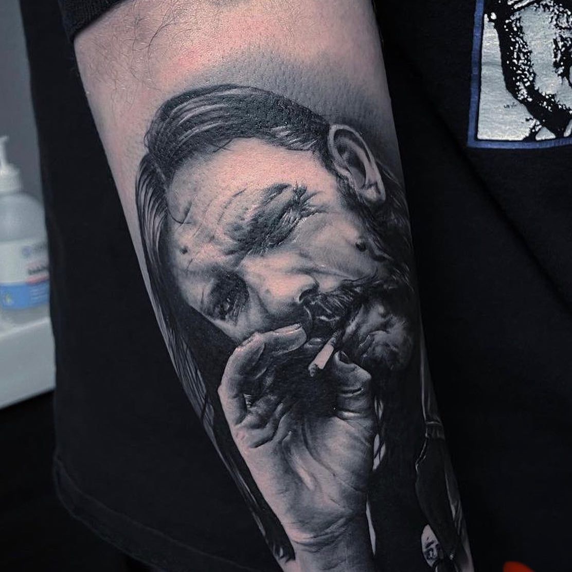 A detailed black-and-grey tattoo on a forearm featuring an angel statue figure with wings above geometric shadow shapes.
