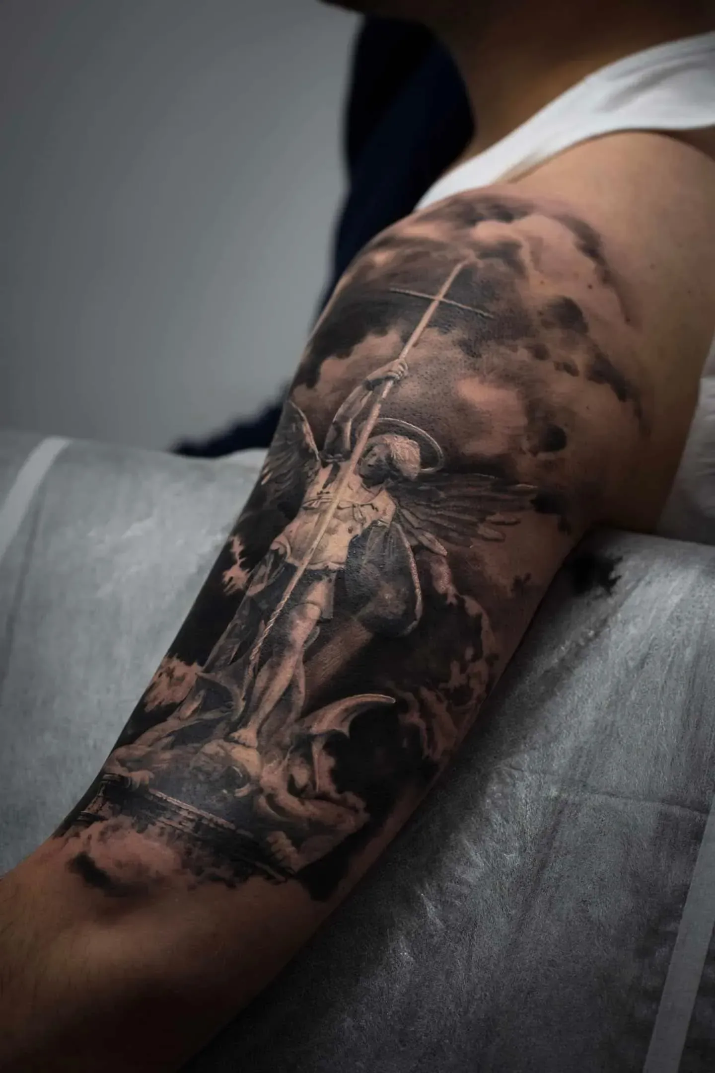 A detailed black-and-grey tattoo on an upper arm depicting an angel subduing a figure amidst dark, swirling clouds.