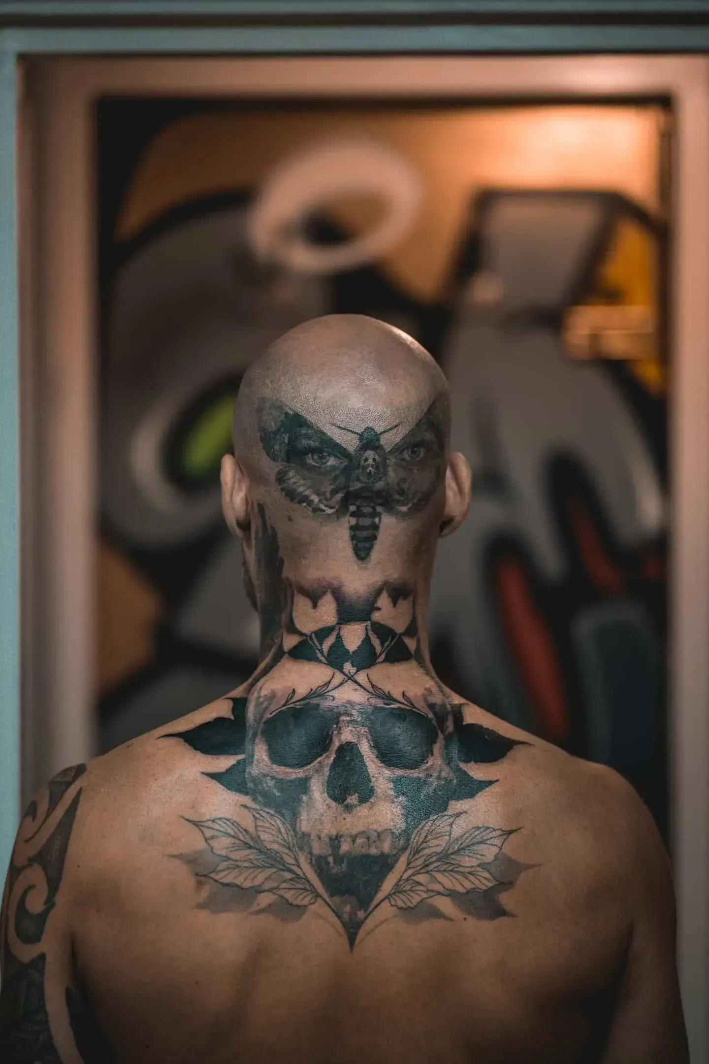 A person seen from behind, displaying a large moth tattoo on the back of their head and a skull design on their upper back.