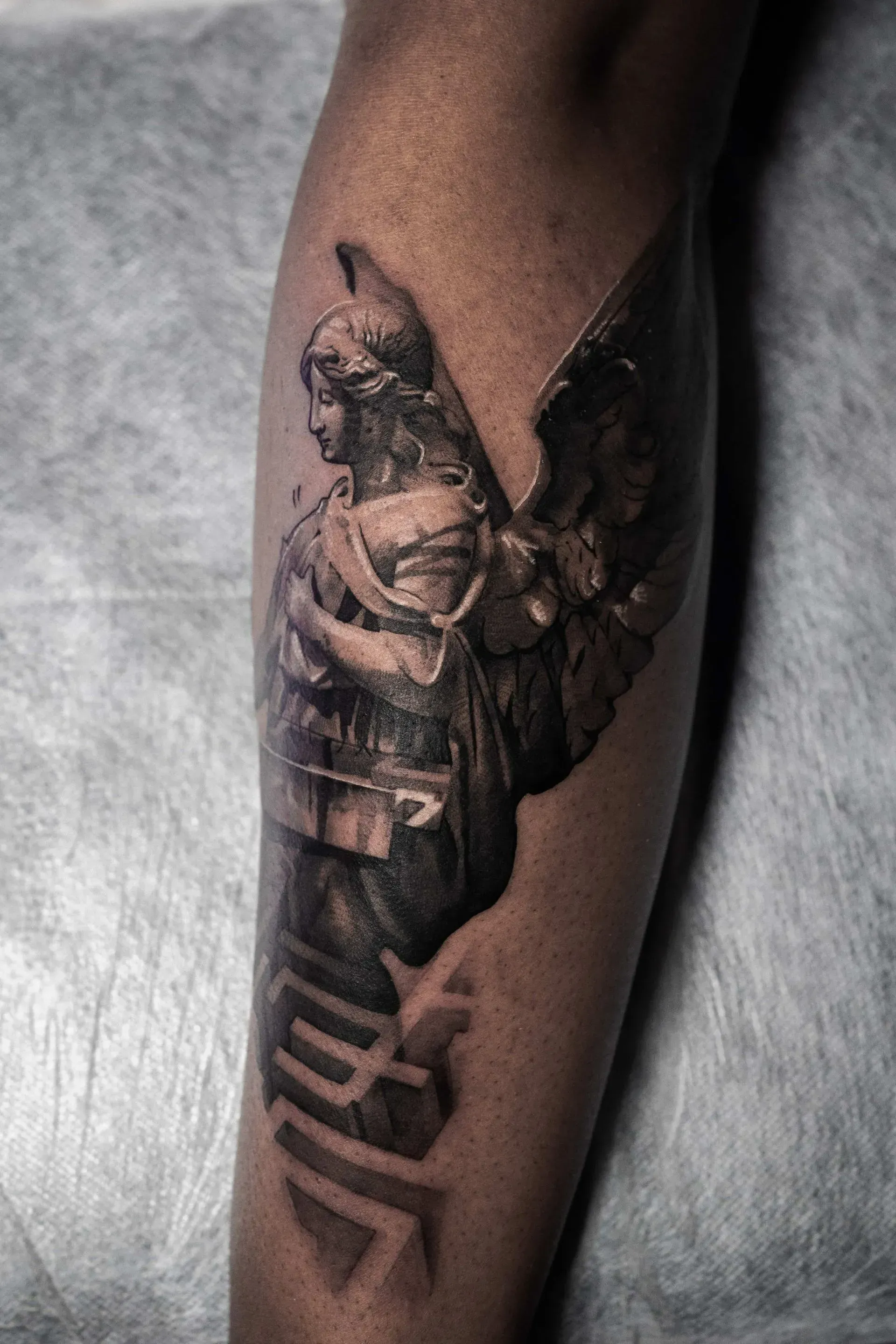 A detailed black-and-grey tattoo on a forearm featuring an angel statue figure with wings above geometric shadow shapes.