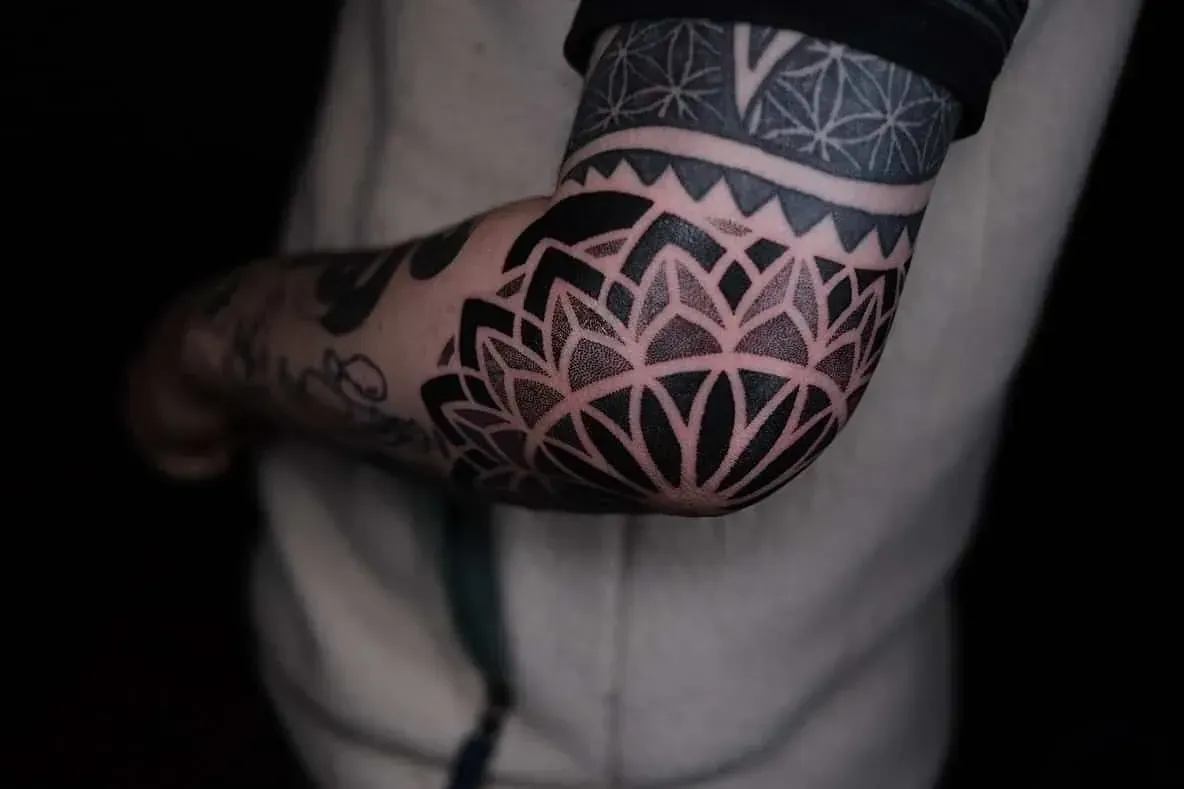 A close-up of a person's elbow featuring a large, black ink mandala tattoo with intricate geometric patterns.