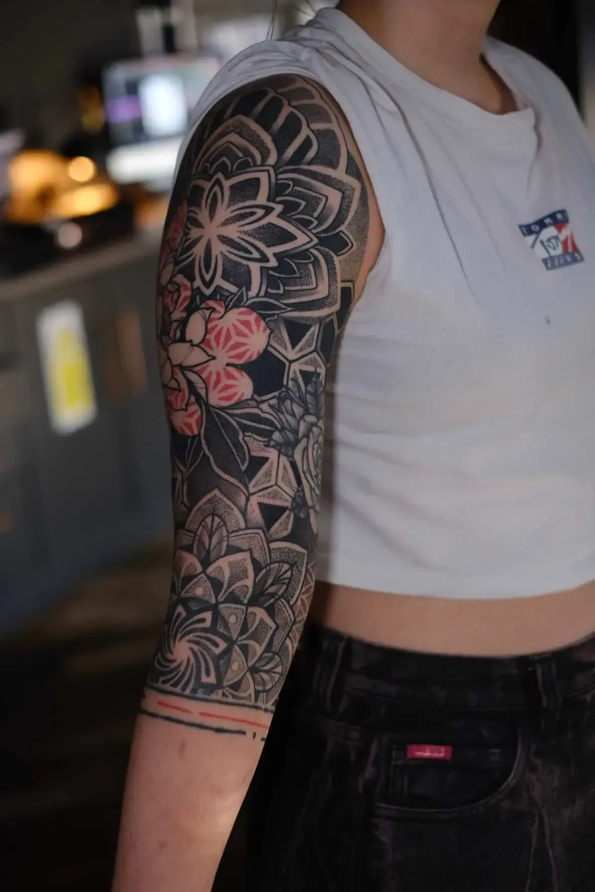 A person’s arm featuring an intricate black-and-grey geometric tattoo sleeve with subtle red accents on a floral element.