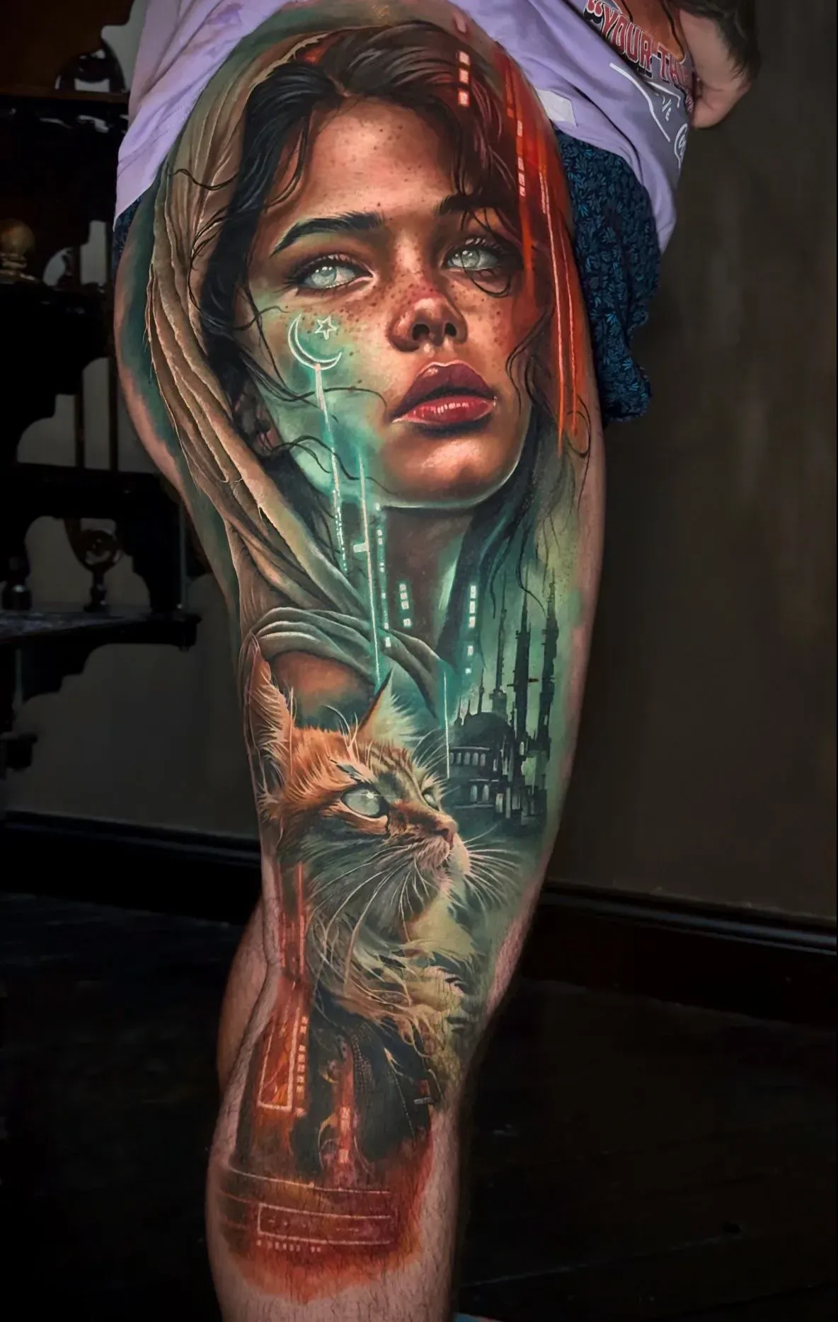 Surreal leg tattoo featuring a glowing-eyed woman with hooded hair above a cat and a silhouetted castle in vibrant colors.