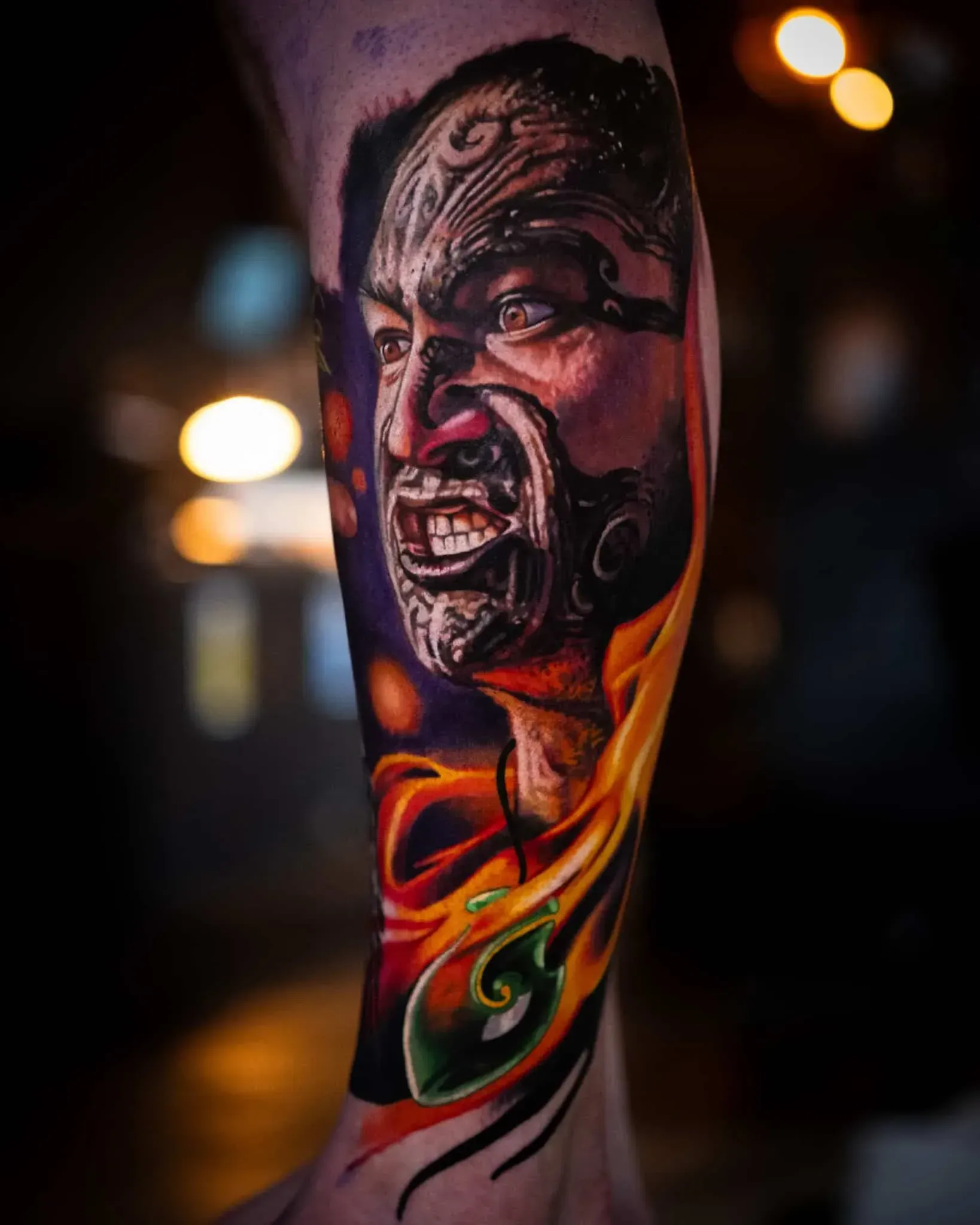 A realistic tattoo on a person's forearm depicting a face with traditional Moko patterns, surrounded by vibrant flames.